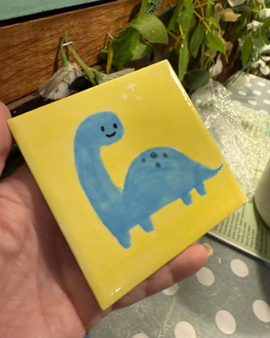 A hand holding a small yellow box with a cute illustration of a blue dinosaur with a happy face on a yellow background.