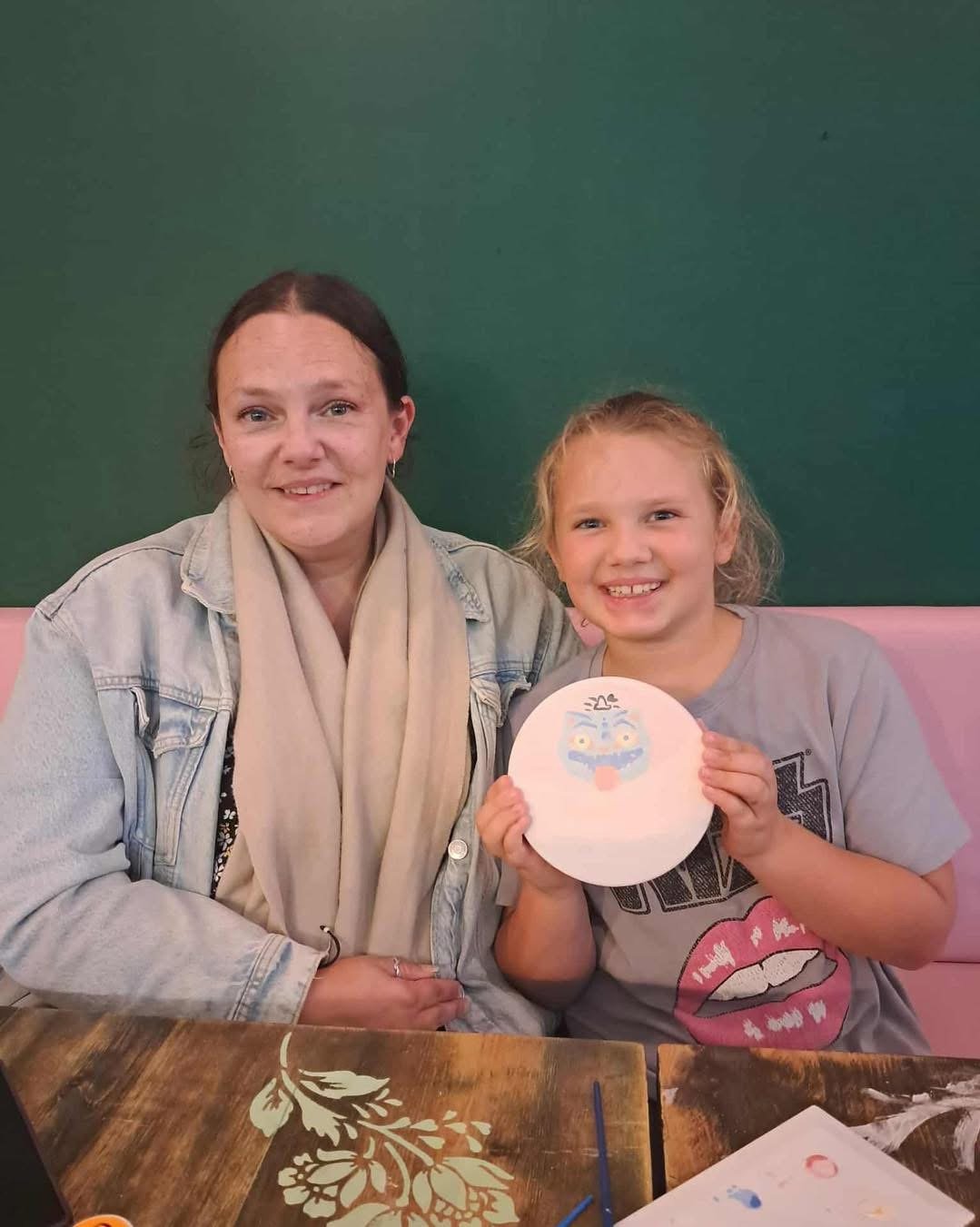 A woman and a young girl sitting together at a table in a restaurant. The girl is holding a circular piece of paper with a colorful face drawn or painted on it, and both are smiling.