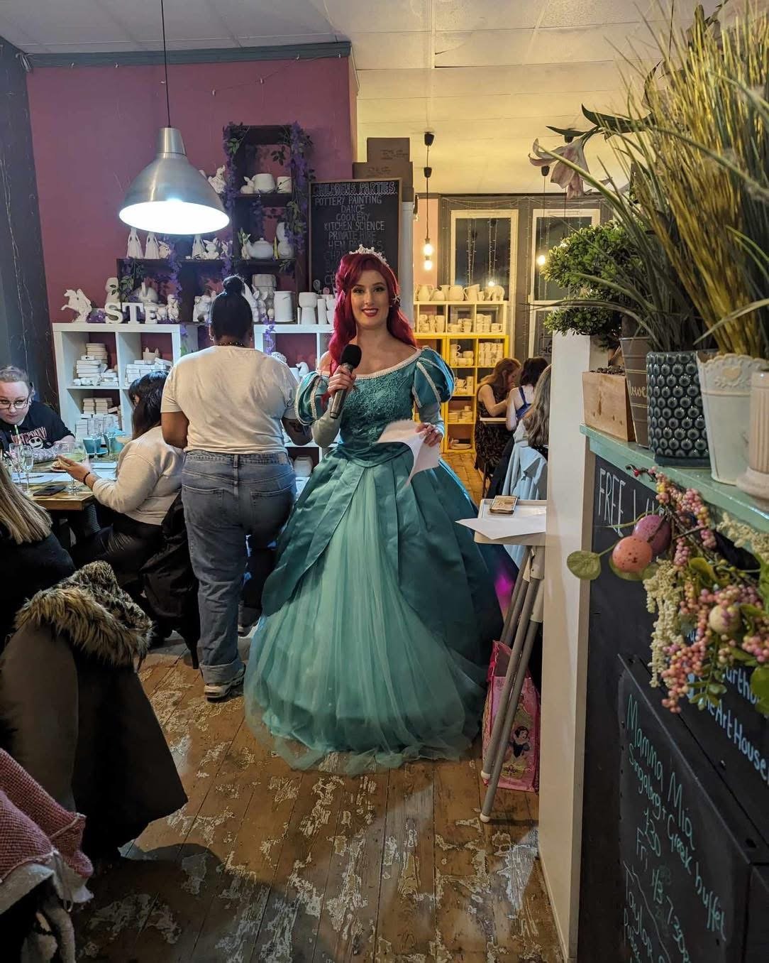 A woman dressed as a princess in a turquoise gown stands in a cozy cafe, holding a microphone and a piece of paper, surrounded by seated patrons and cozy decor.
