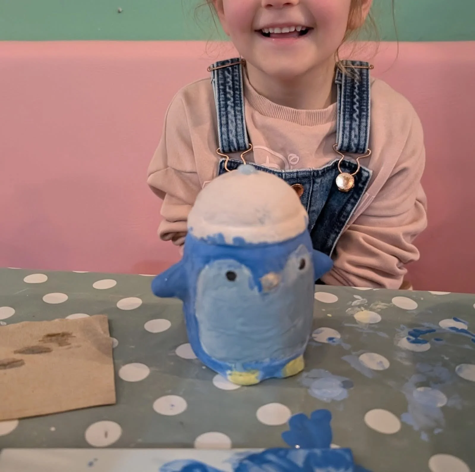 Close-up of a young girl smiling and showing missing teeth, wearing denim overalls and a beige long-sleeve shirt, sitting at a table with a painted ceramic penguin in front of her, and a polka dot tablecloth in the background.