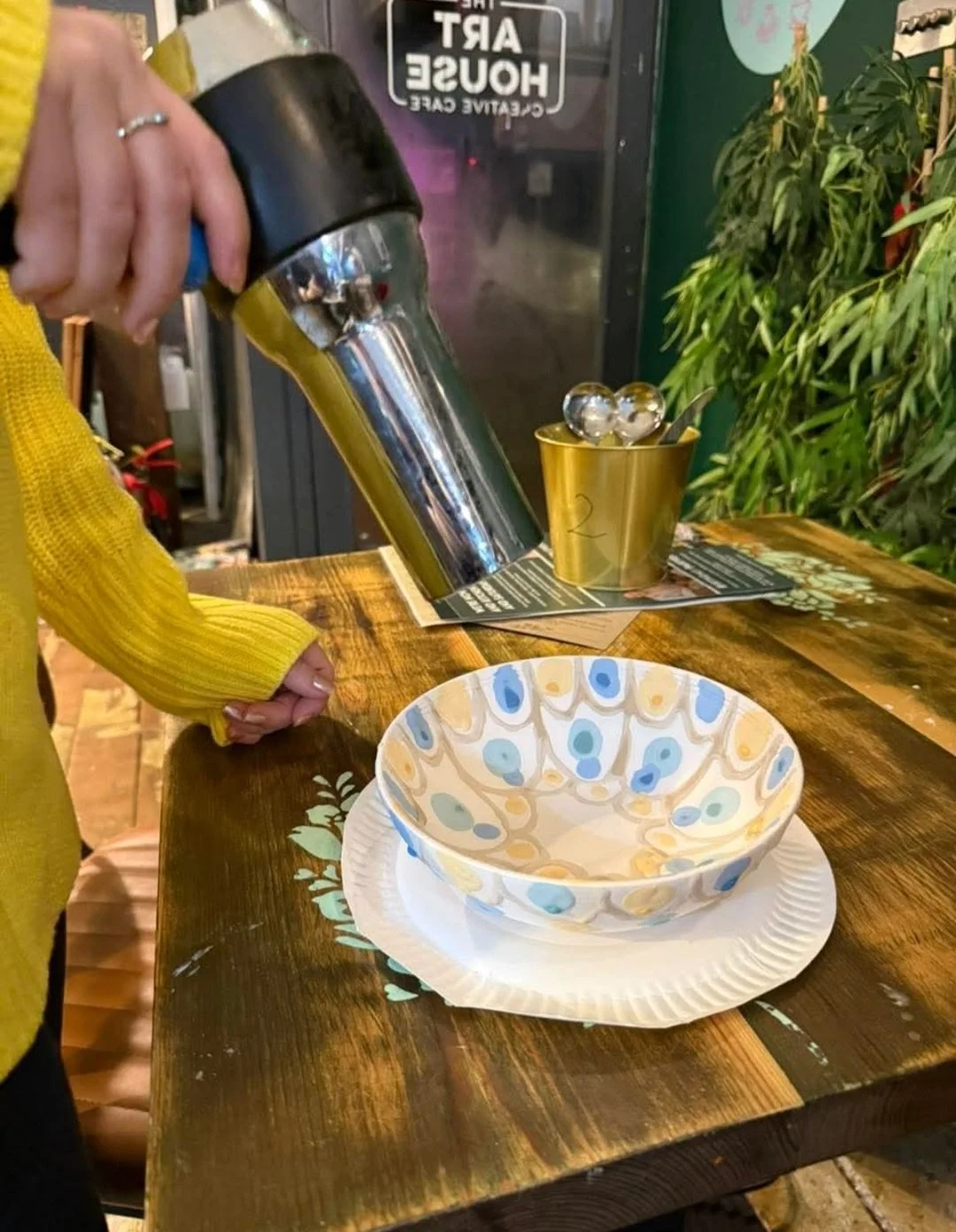 A person wearing a yellow sweater is pouring honey from a squeeze bottle into a decorated bowl placed on a paper plate on a wooden table, near a gold-colored container holding spoons and balls.