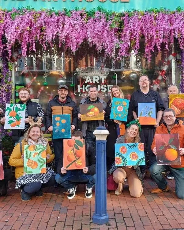 Group of people holding colorful paintings of fruit and nature scenes outside a cafe decorated with hanging pink flowers.