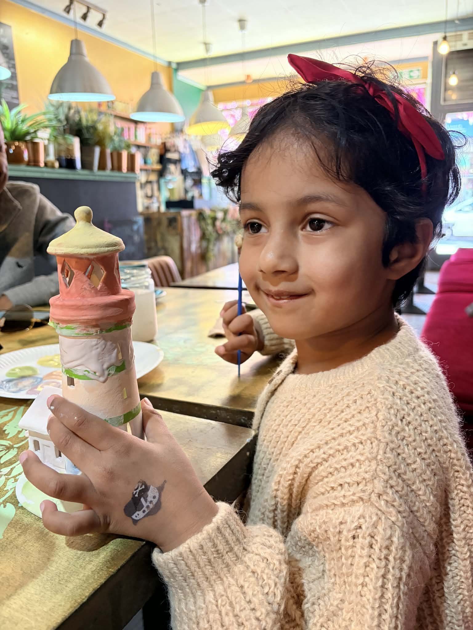 A young girl with short black hair and a red headband, wearing a beige knit sweater, is sitting at a table in a brightly colored cafe. She is holding a decorated ice cream cone with pink, yellow, and green icing and a castle-like design. She has a small tattoo of a horse on her hand.