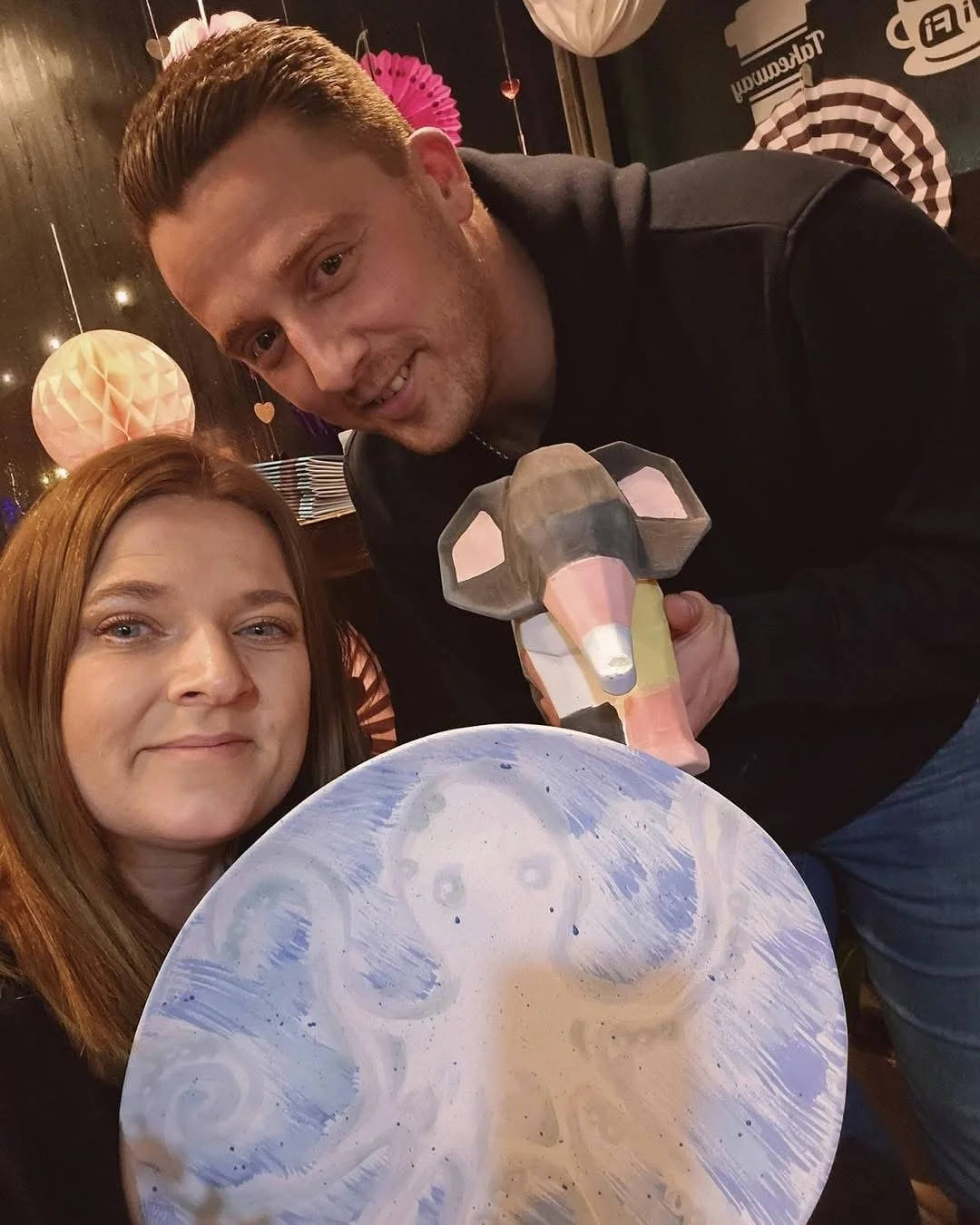 A man and a woman taking a selfie in a decorated indoor setting. The woman holds a round, swirled, celestial-themed art piece in front of her. The man is holding a colorful, geometric elephant sculpture.