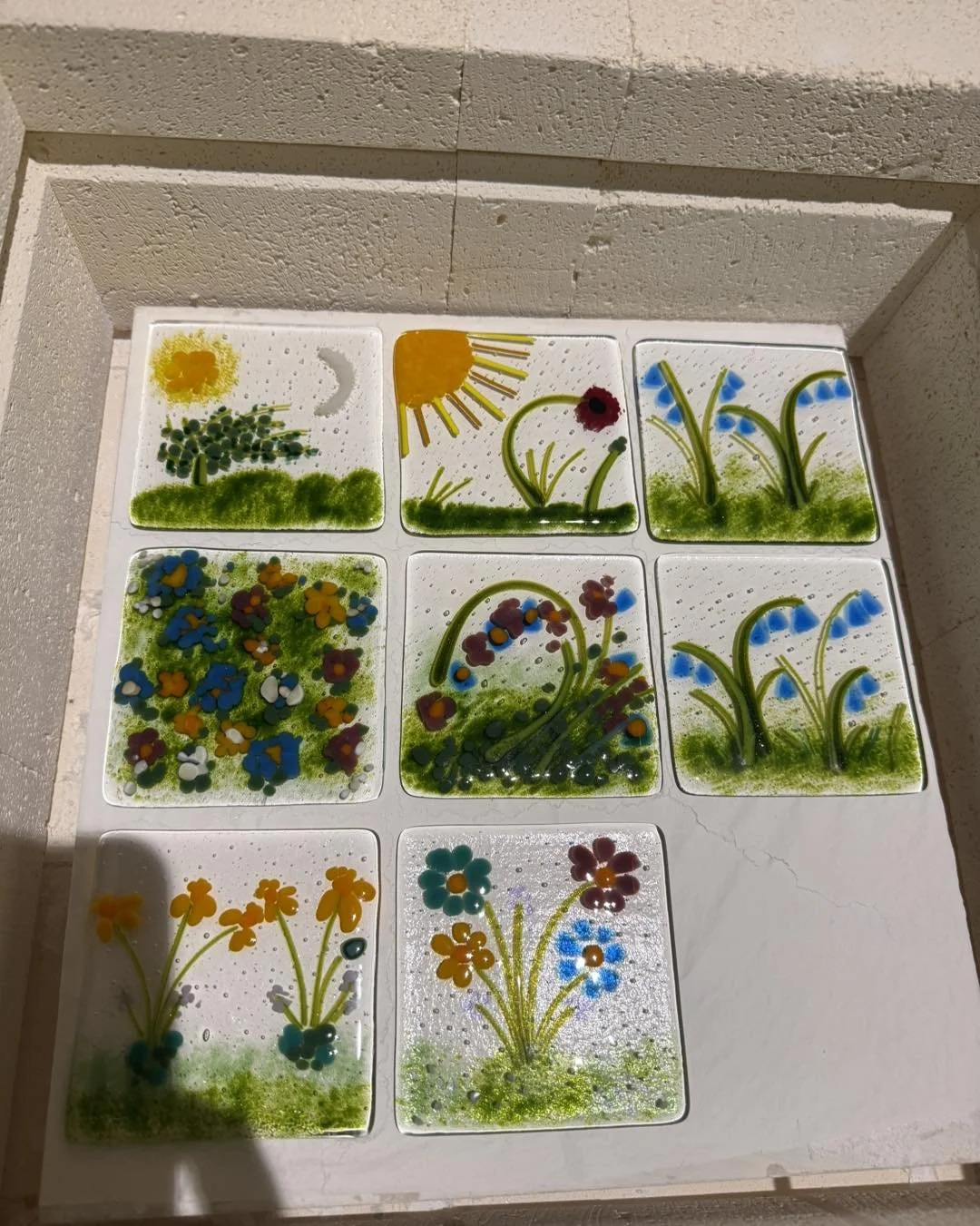 Decorative glass tiles with colorful floral and nature designs, including flowers, sun, moon, and grass, displayed on a white surface.