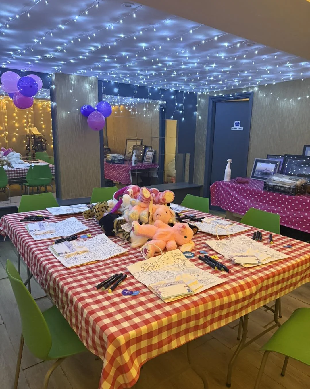 A decorated room with fairy lights on the ceiling, purple balloons, toys and coloring pages on a table with a red and white checkered tablecloth, and green chairs around it, indicating a party or children's event.