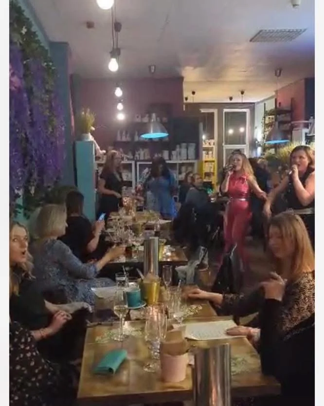 A lively indoor restaurant or cafe scene with people socializing, eating, and drinking. Several women are seated at a long table, some using their phones, while others chat or enjoy beverages. In the background, a few women are standing and talking. The decor features bookshelves, plants, and hanging lights, creating a cozy, inviting atmosphere.