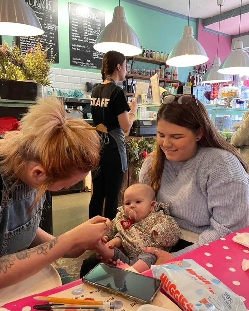 A woman holding a baby in a cafe with another woman looking at the baby. A staff member stands in the background behind the counter, with menu boards and shelves behind her. The table has a pink polka dot tablecloth, a phone, and some papers.