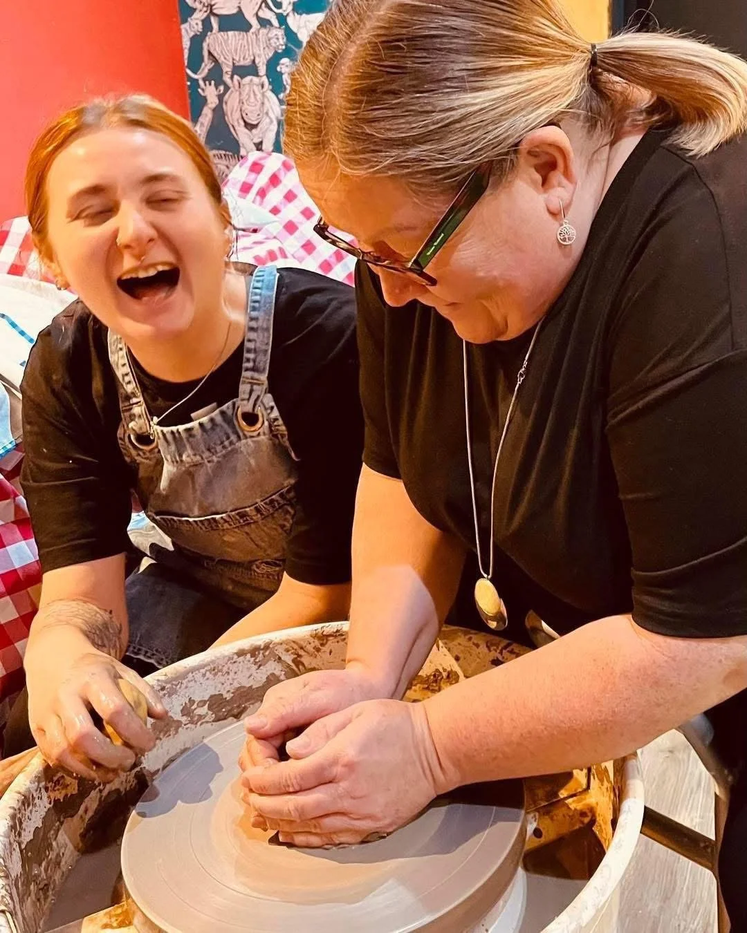 Two women happily working together on a pottery wheel. One woman wearing glasses and a black shirt is shaping clay, while the other woman with short hair and a black shirt laughs joyfully. The background features colorful decor and a mural.