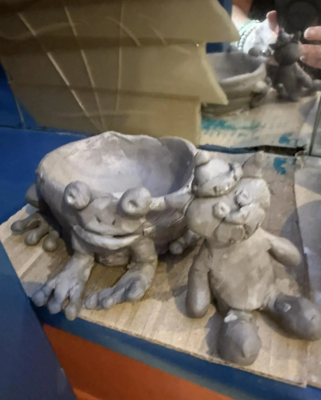 Unfinished clay sculpture of a crab holding a bowl on a wooden surface.