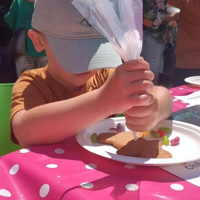 A young boy wearing a gray baseball cap and an orange shirt is squeezing pink frosting onto decorated cookies on a white plate. The table has a pink polka dot tablecloth, and there are other people and plates in the background.