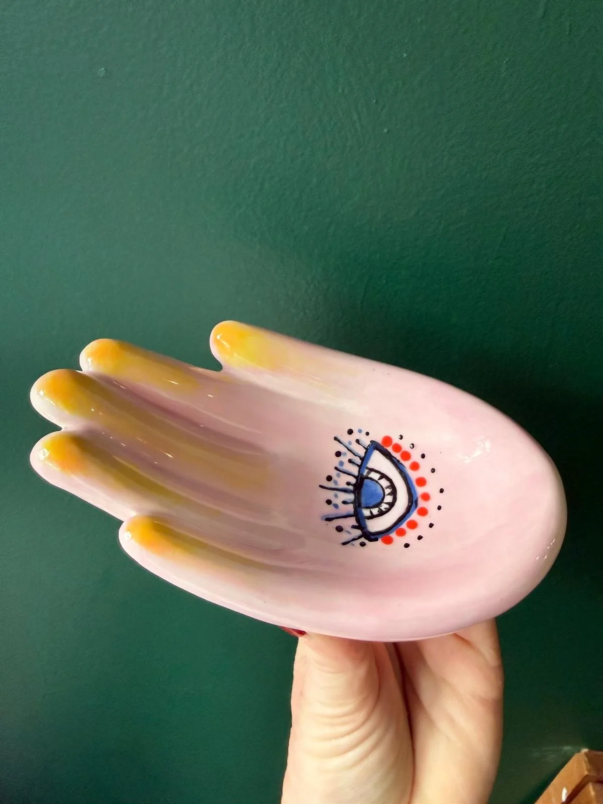 Ceramic hand-shaped dish with eye and face design, held by a person against a green background.