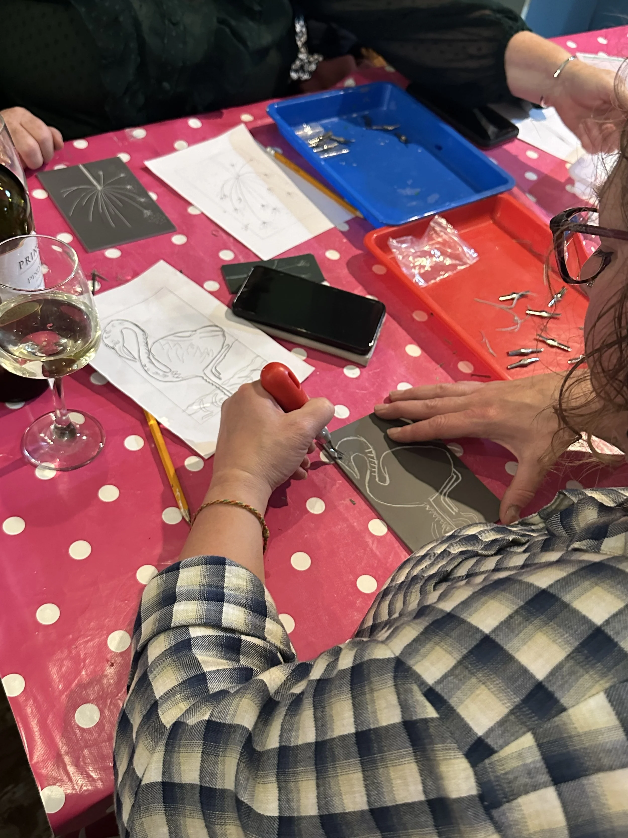 People working on a craft project at a table covered with a pink and white polka dot tablecloth. There are papers with designs, small scissors, and plastic bags on the table. One person's hand is holding a tool, working on a black piece of material with a flamingo design.