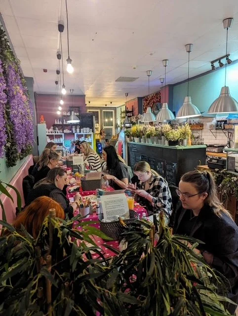 People sitting at a pink table in a cozy cafe with floral and plant decor, working or chatting.