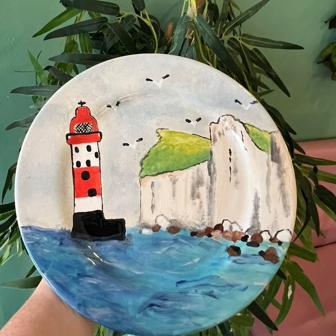Decorative ceramic plate painted with a lighthouse, white cliffs, and the ocean, surrounded by green plant leaves.