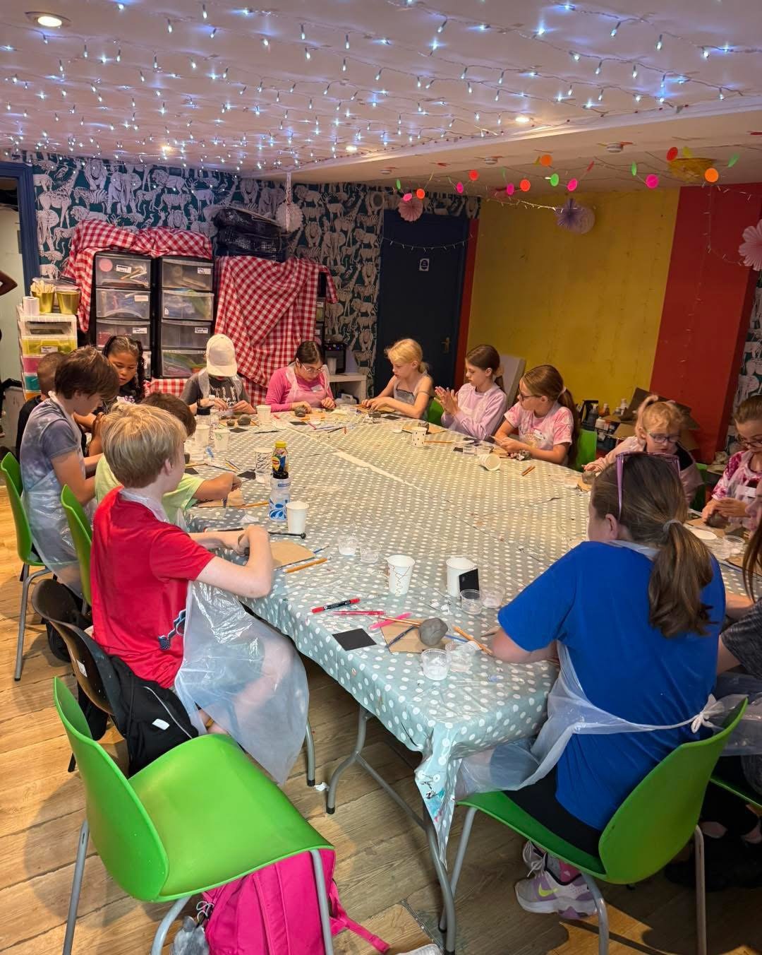 Children participating in a craft activity around a large, polka-dotted table in a decorated room with string lights and colorful wall decorations.