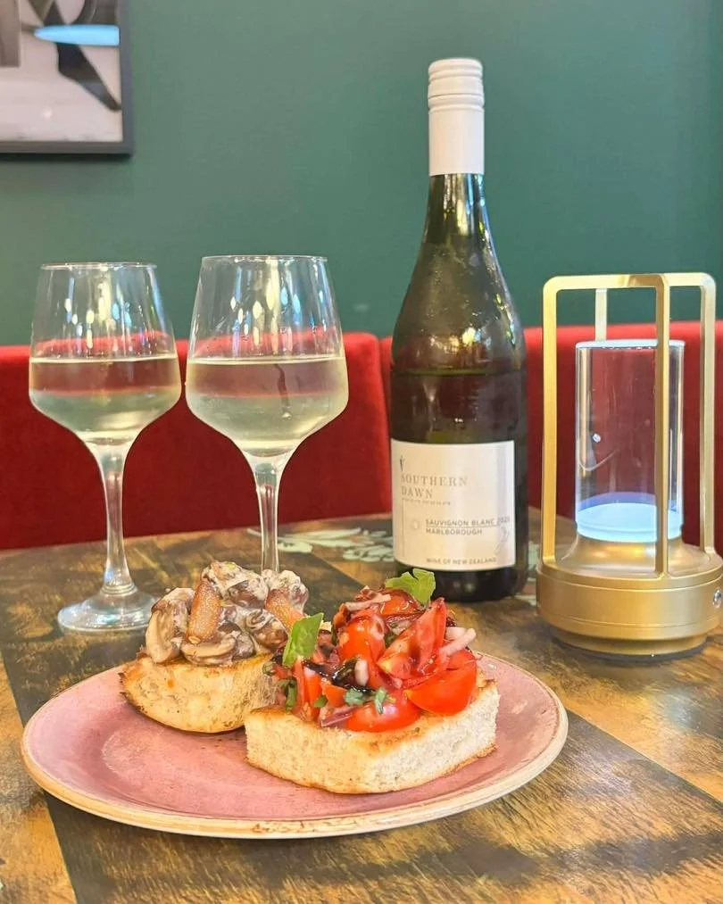 Two glasses of white wine, a bottle of wine, a plate with bruschetta topped with tomato and herbs, and an empty gold and glass lantern on a wooden table in a restaurant setting.