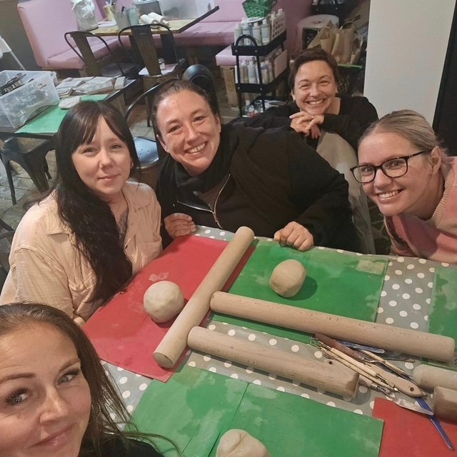 Group of five women sitting around a table with rolling pins and dough, smiling at the camera in a kitchen or bakery setting.