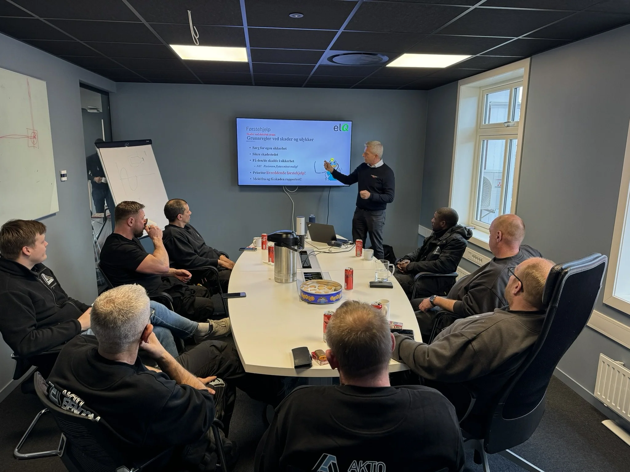 En grupp personer deltar på et foredrag eller møte i et konferanserom. En person står foran en skjerm og snakker til publikum, mens andre sitter rundt et langbord, og noen har drikkevarer og snacks foran seg.