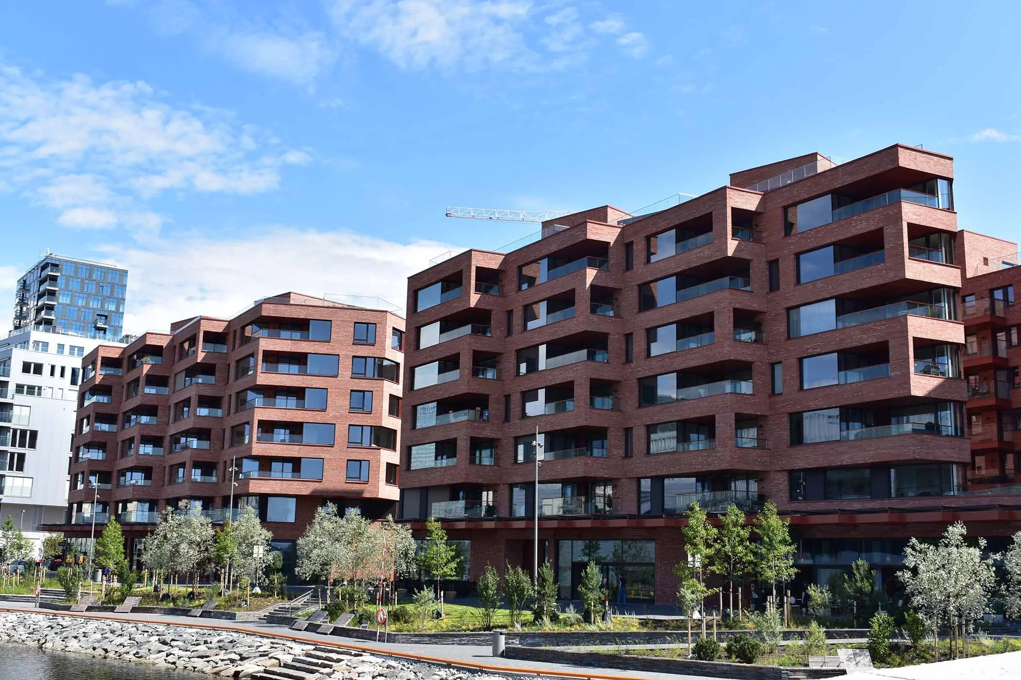 Fleretasjet leilighetsbygg i murstein, med store vinduer og balkonger, omringet av grøntområder og et vann med steiner i forgrunnen mot en blå himmel med spredte skyer.