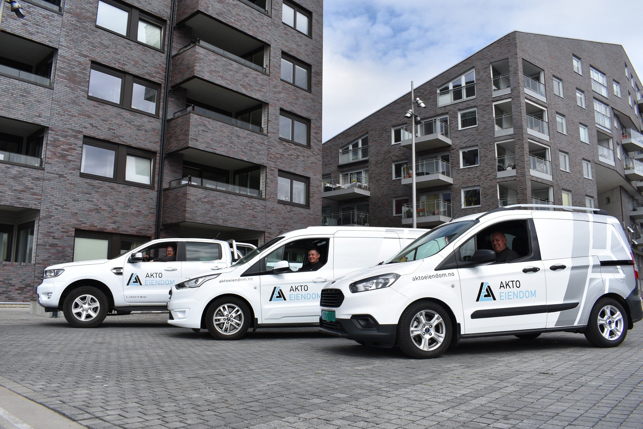Tre hvite firmabiler med logo og nettsideadresse kjører foran et modern bygg med balkonger. To personer sitter i bilene og smiler.