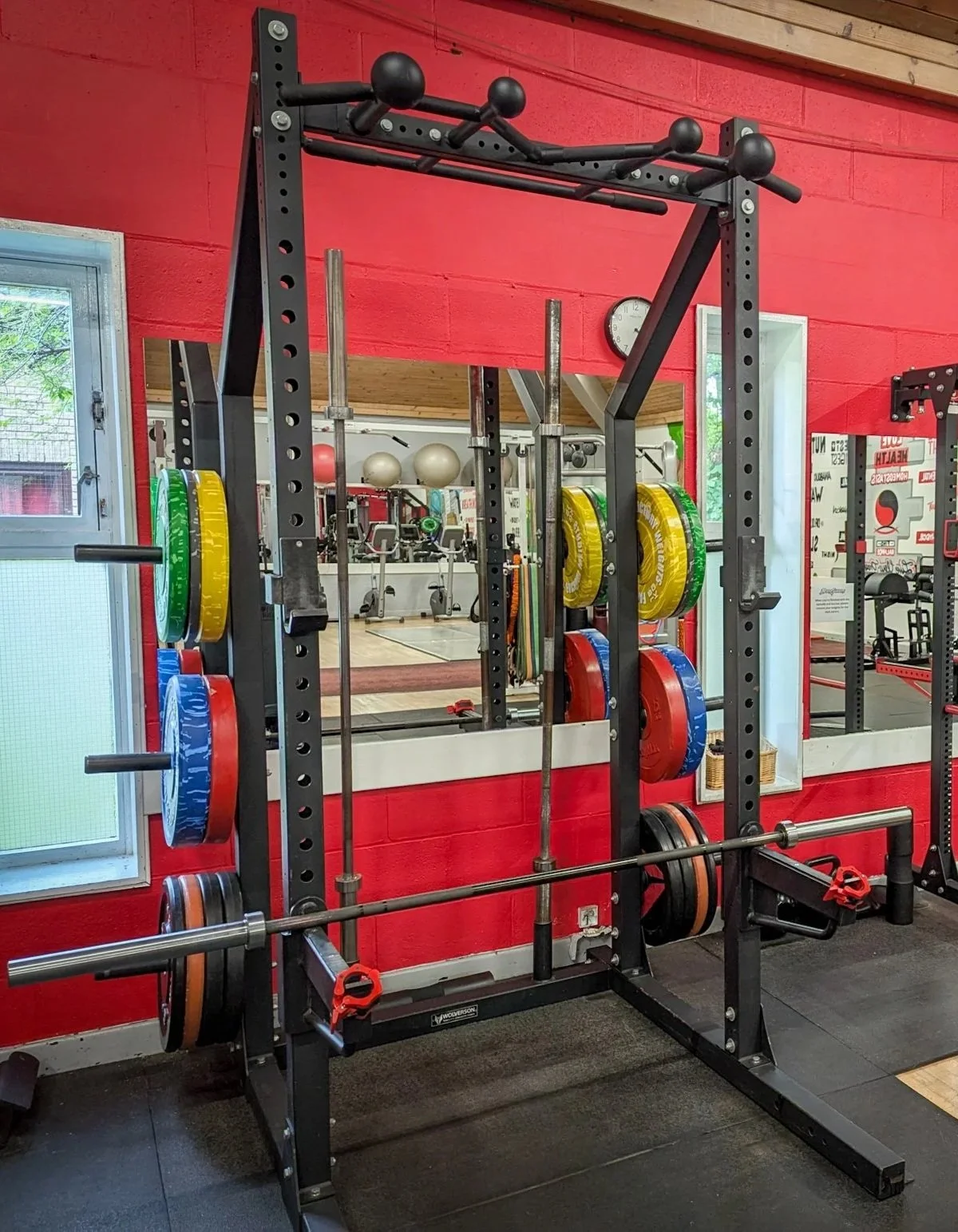 A weightlifting power rack with colorful weight plates, a barbell, and a mirror reflecting gym equipment in the background, with red walls and a clock.