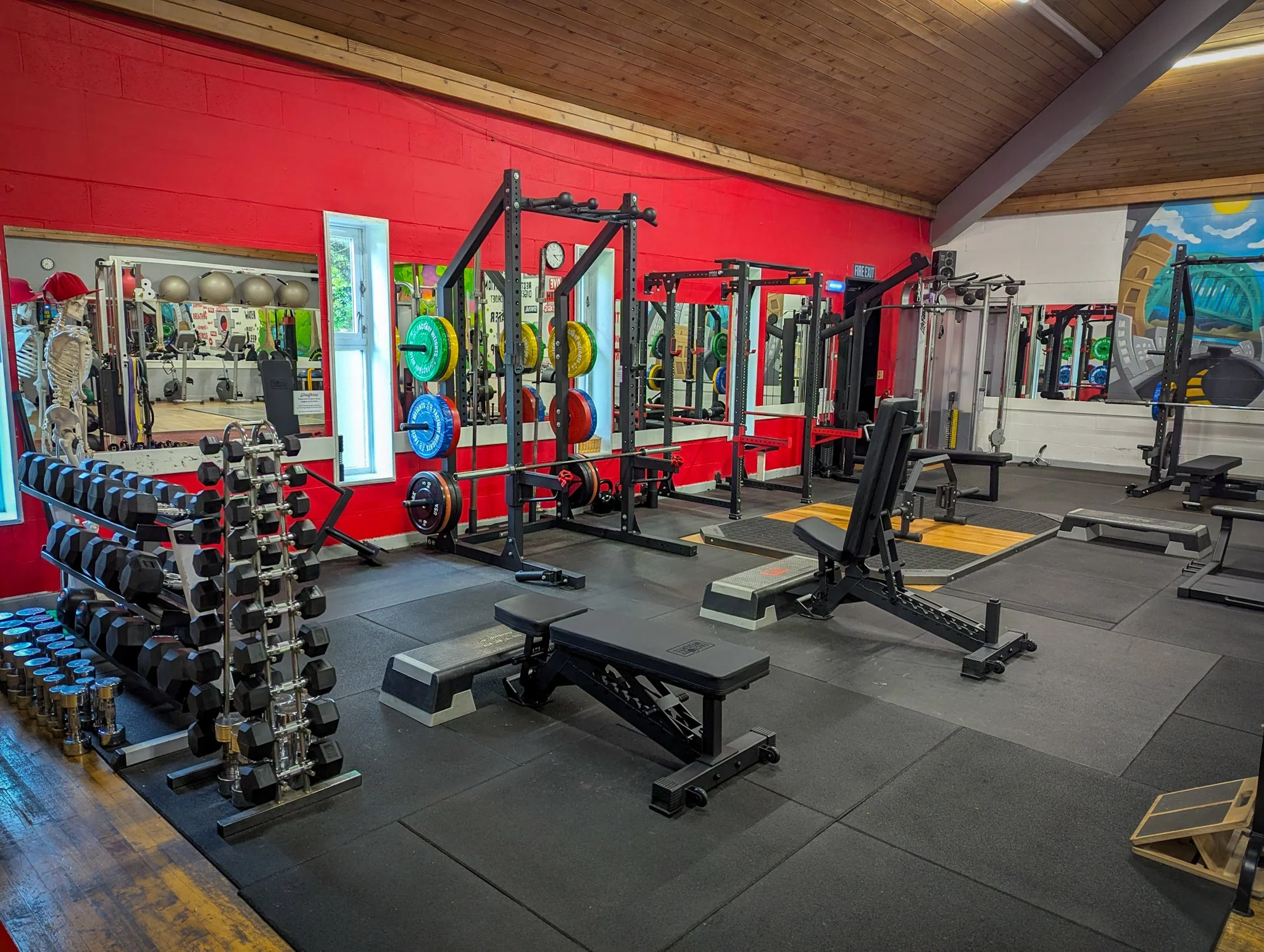 View of a gym with free weights, weightlifting equipment, a mirror, and a colorful mural on the wall.