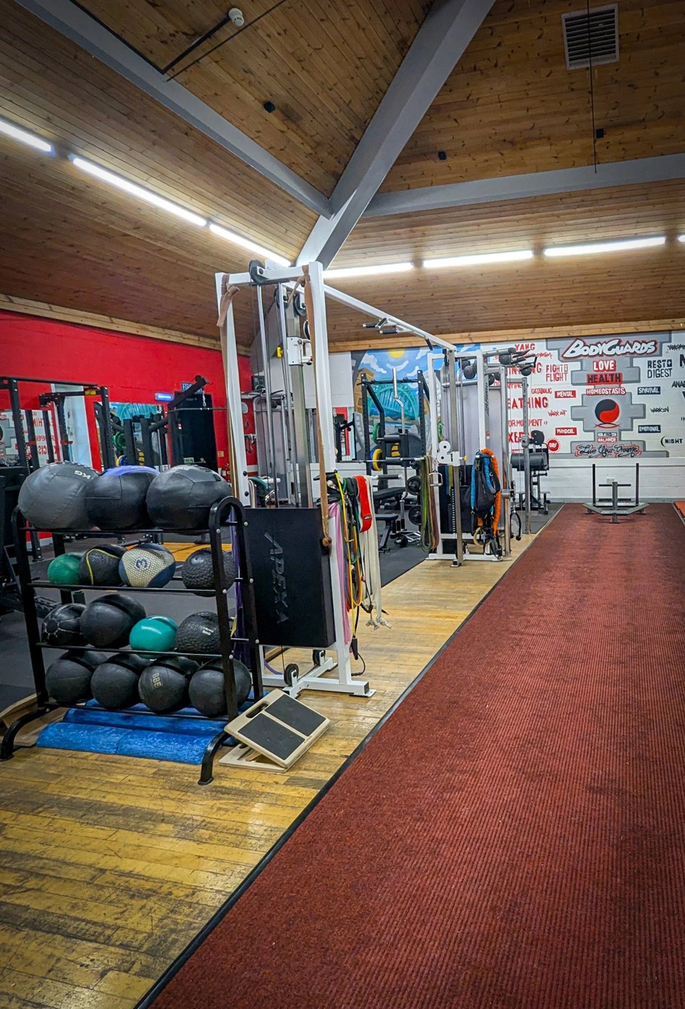 Indoor gym with various equipment including kettlebells, medicine balls, resistance bands, weightlifting machines, and a red carpeted area on wooden flooring, with a wall decorated with motivational words and graphics.
