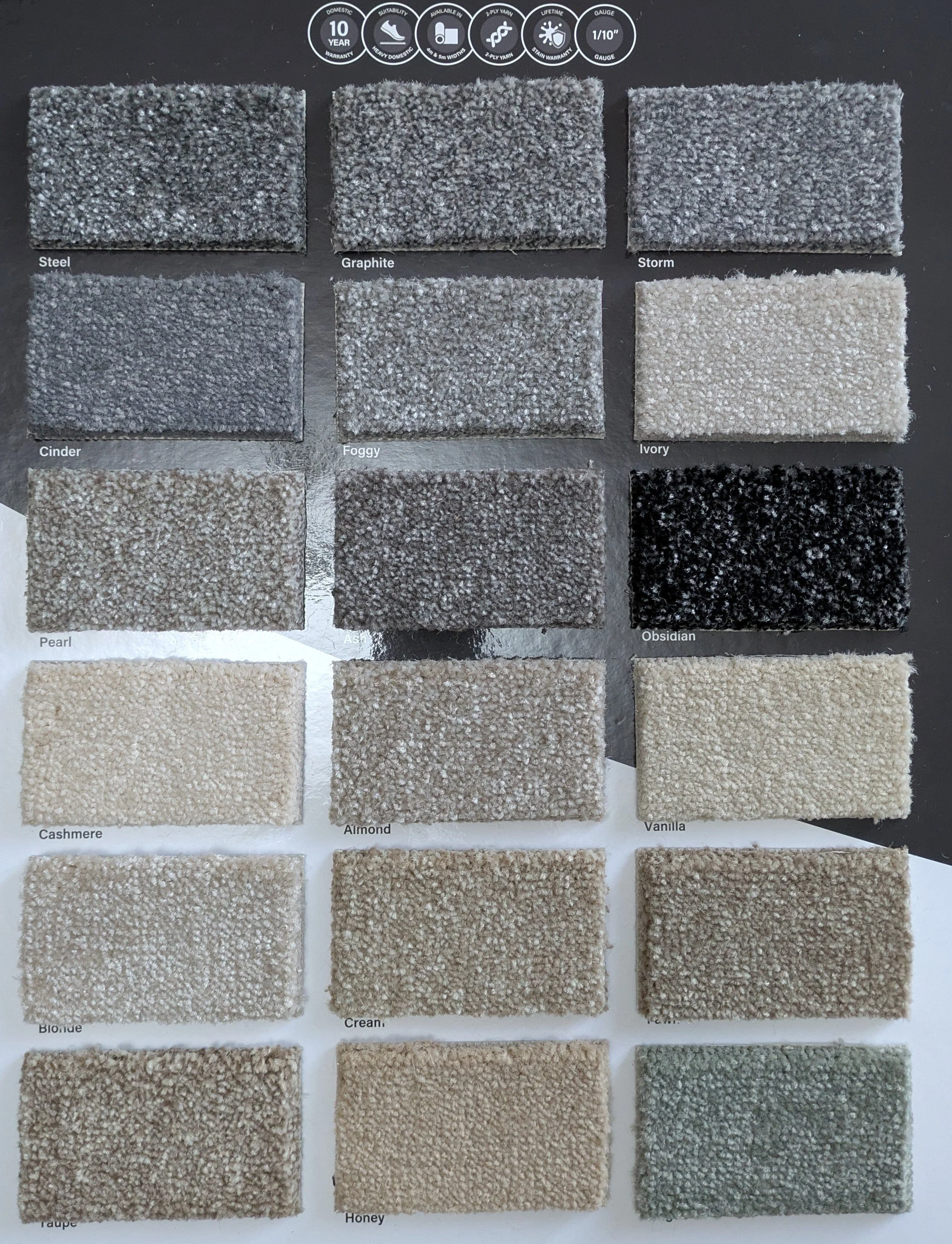 A carpet sample display with 12 different shades of neutral colors arranged in four rows and three columns. The shades are labeled as Steel, Graphite, Storm, Cinder, Foggy, Ivory, Pearl, Ash, Obsidian, Taupe, Almond, and Vanilla.