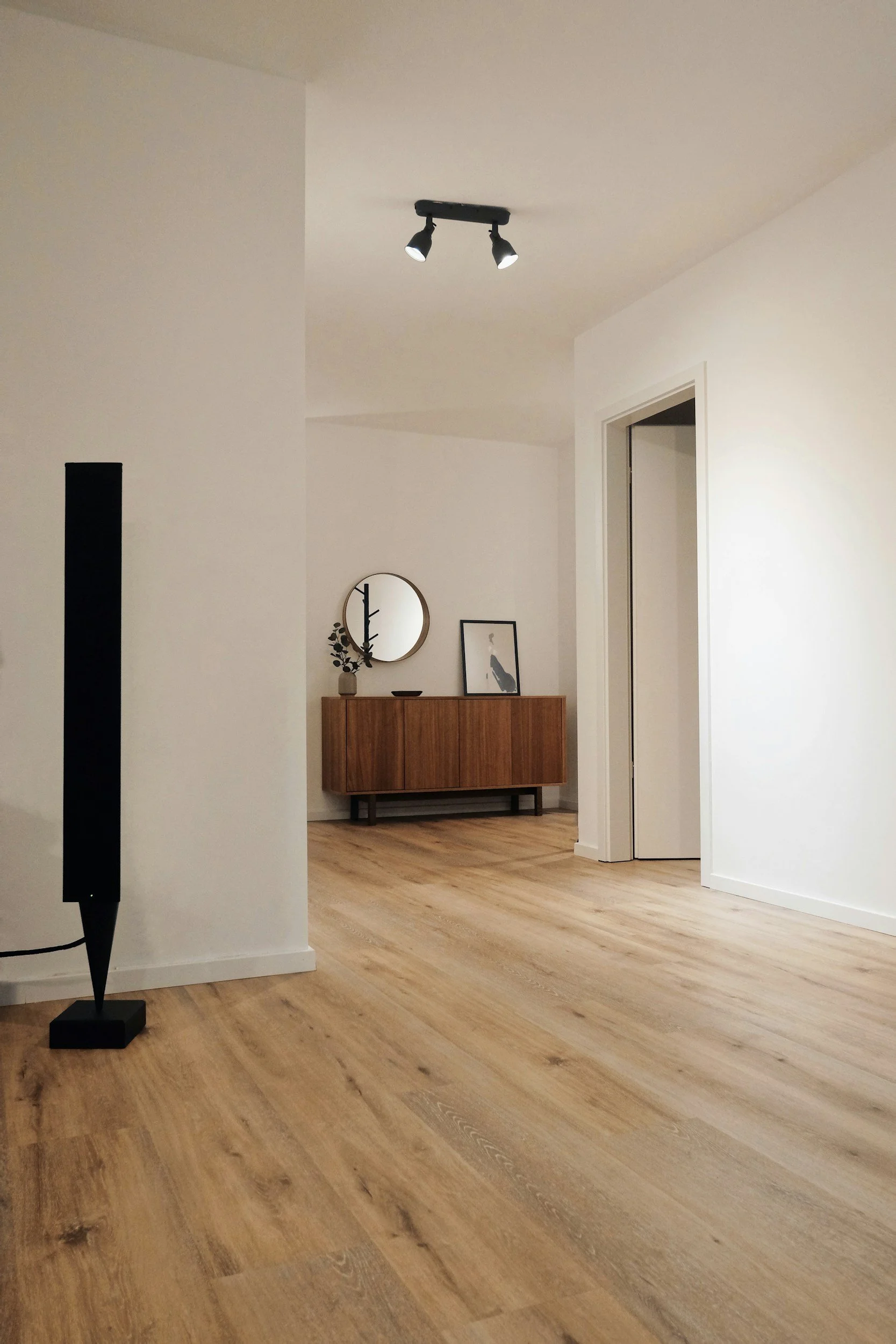 A modern, minimalistic living room with light wood flooring, white walls, and a doorway. A wooden sideboard with framed artwork and a round mirror hang above it. A tall black floor lamp is positioned to the left.