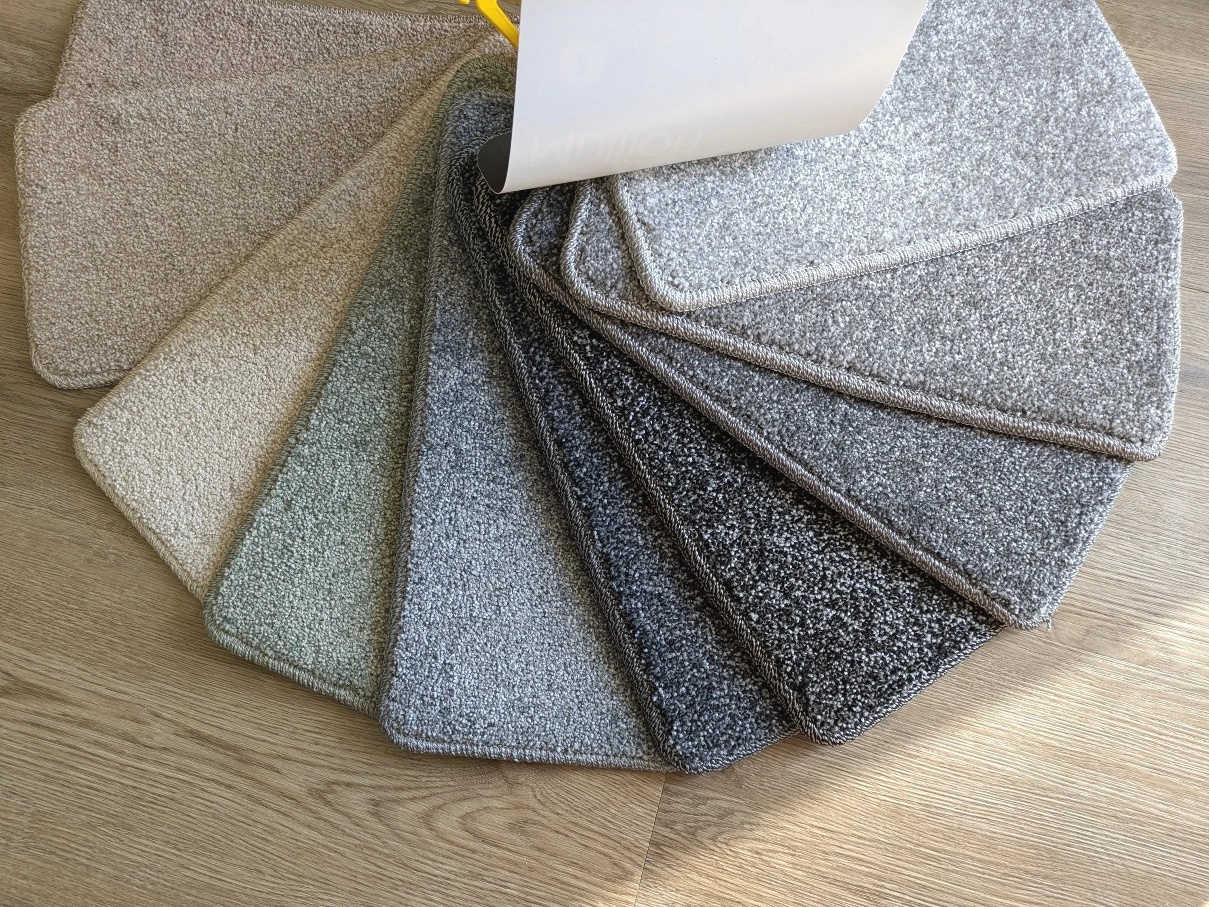 A selection of carpet samples in various shades of grey, beige, green and blue, layered on a wooden floor.