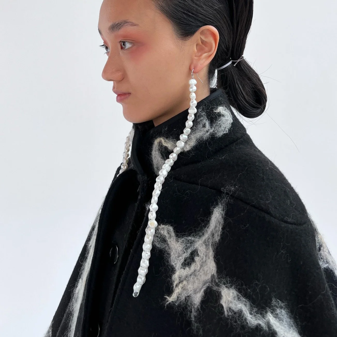 A woman with dark hair styled in a low bun, wearing pearl earrings and a black coat with white fuzzy accents, standing in profile against a plain white background.