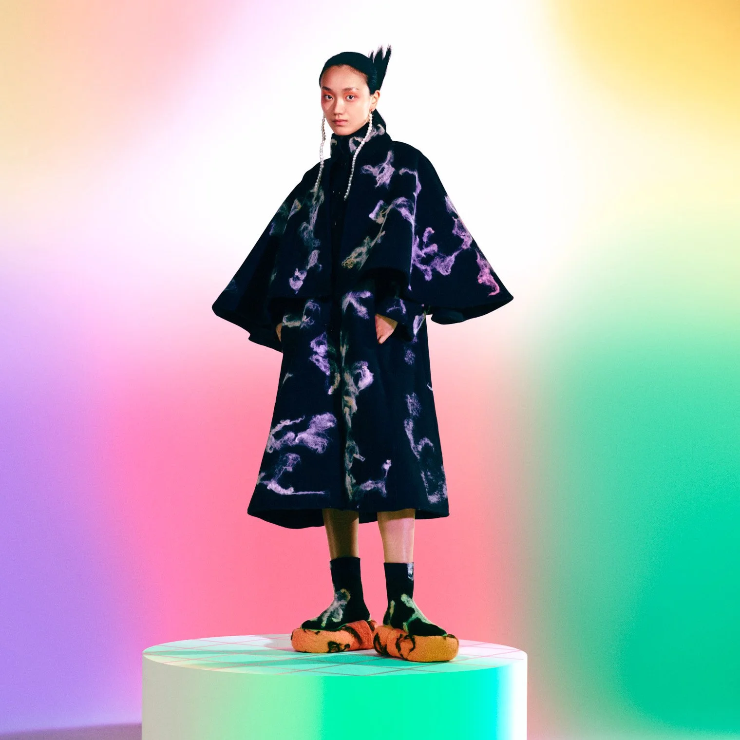 A woman standing on a white circular platform against a colorful, rainbow gradient background. She is wearing a black coat with purple and gold cloud-like patterns, black socks with similar designs, and orange slippers. She has long earrings, and her black hair is styled with peaks.