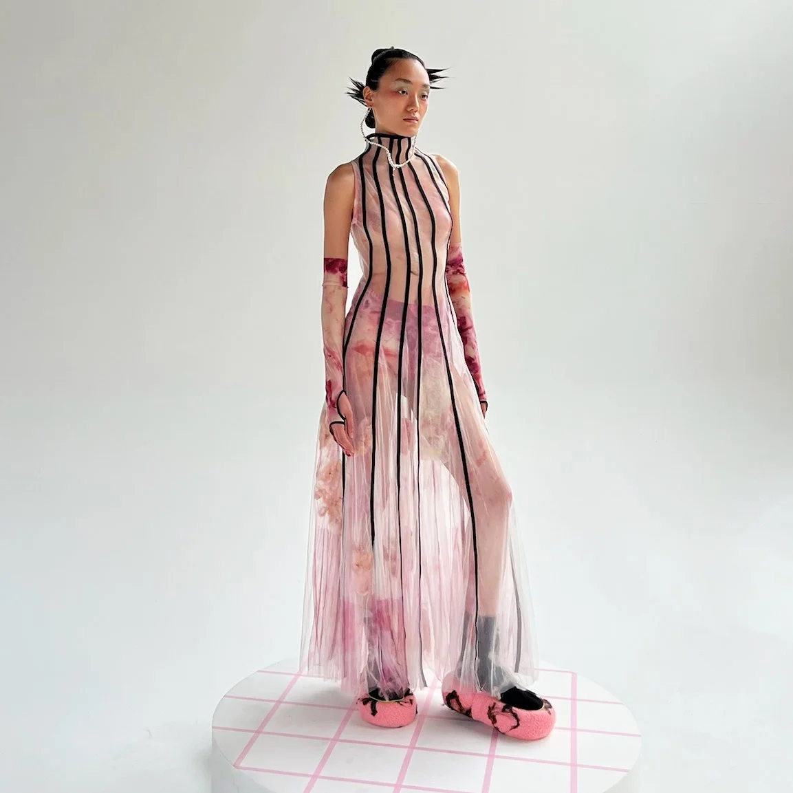 A female model wearing a sheer, sleeveless, pink dress with black vertical stripes, layered over a floral print outfit. She has sheer long gloves matching the dress, black shoes with pink fluffy slippers, and styled hair. She stands on a white platform with a grid pattern.