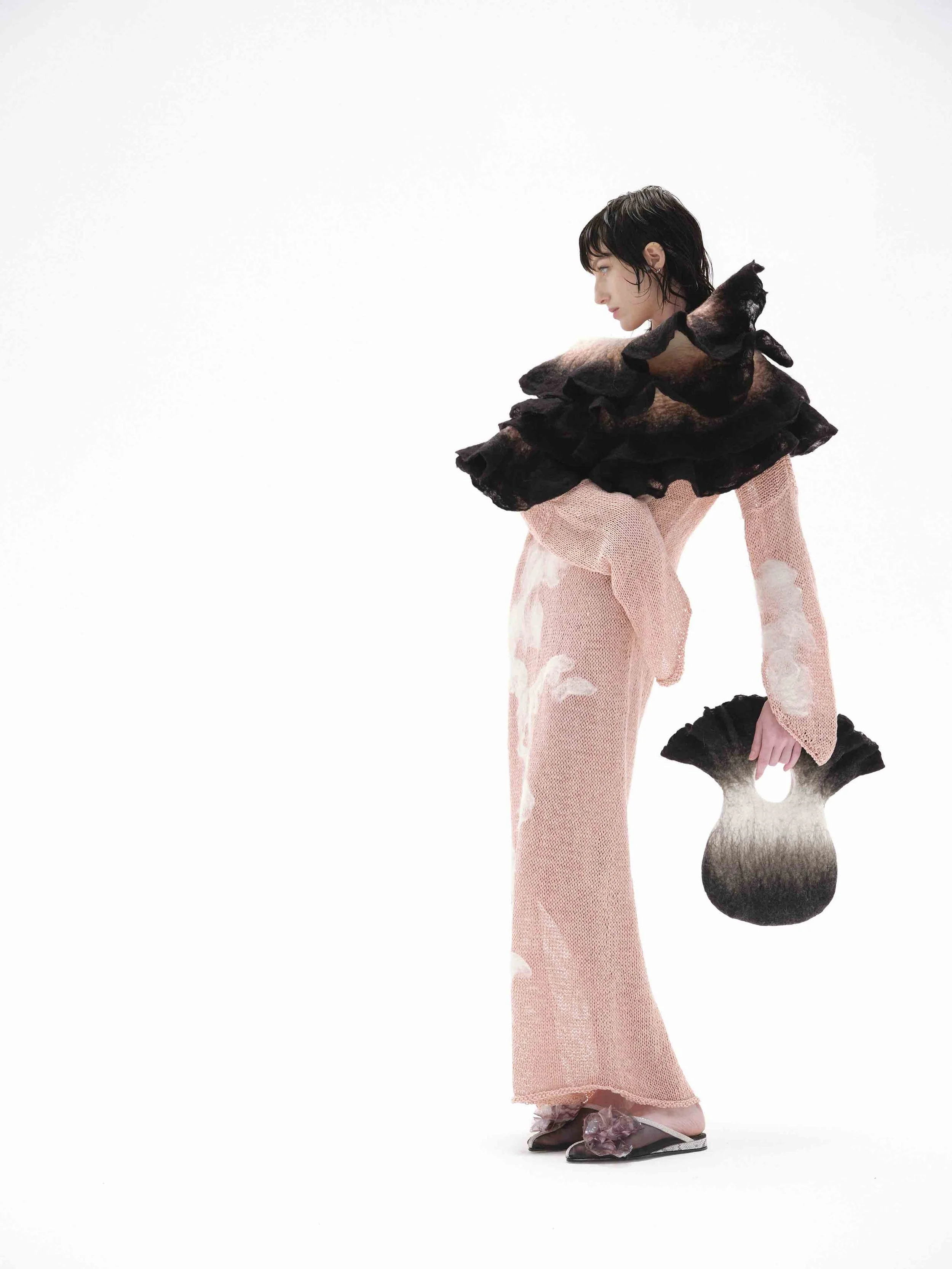 Person in pink textured outfit with wide ruffled collar holding a unique, fan-shaped black and white accessory against a white background.