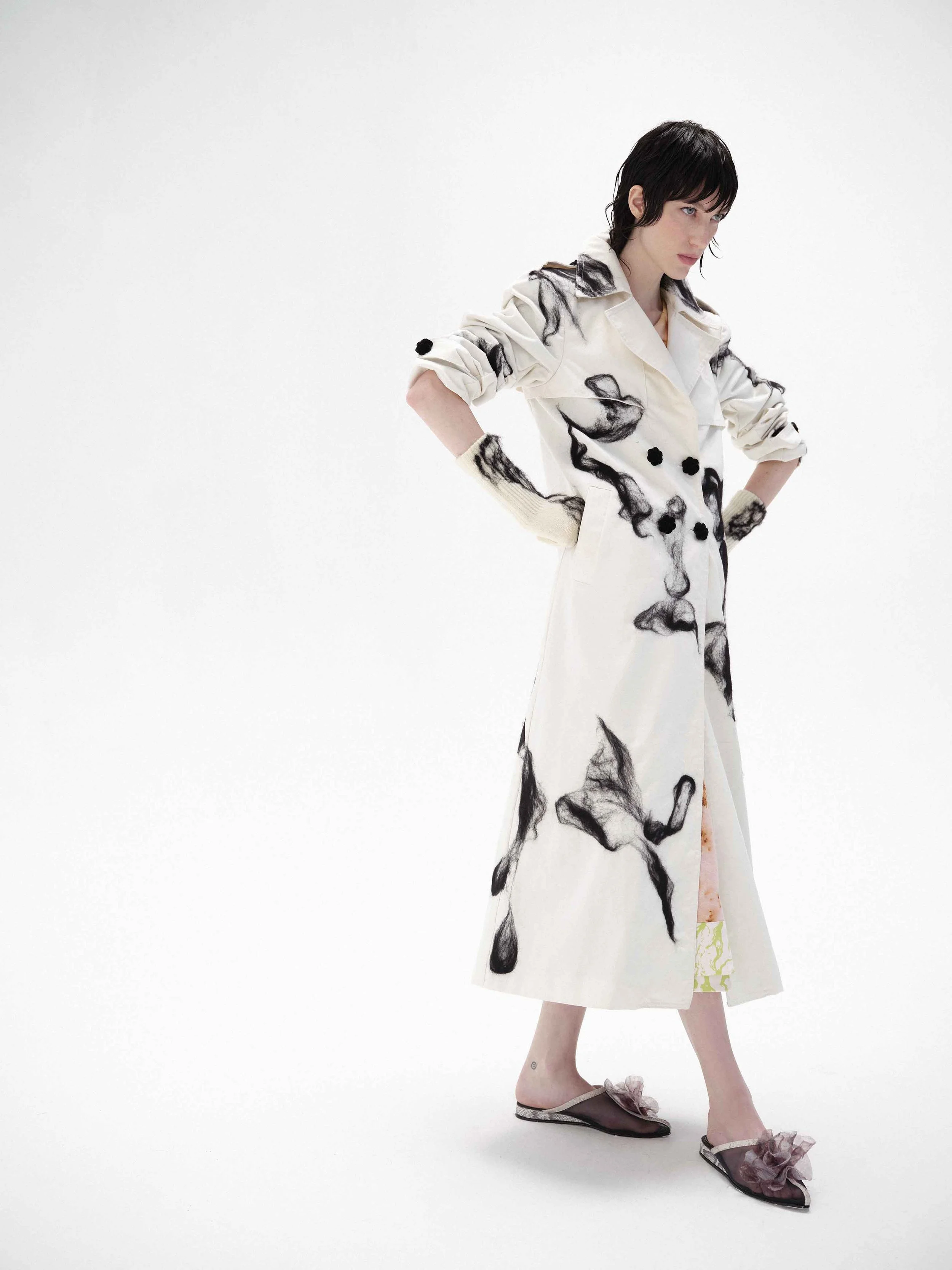 Fashion model wearing a long white coat with black abstract patterns, black hair, and open-toed shoes with decorative flowers on a white background.