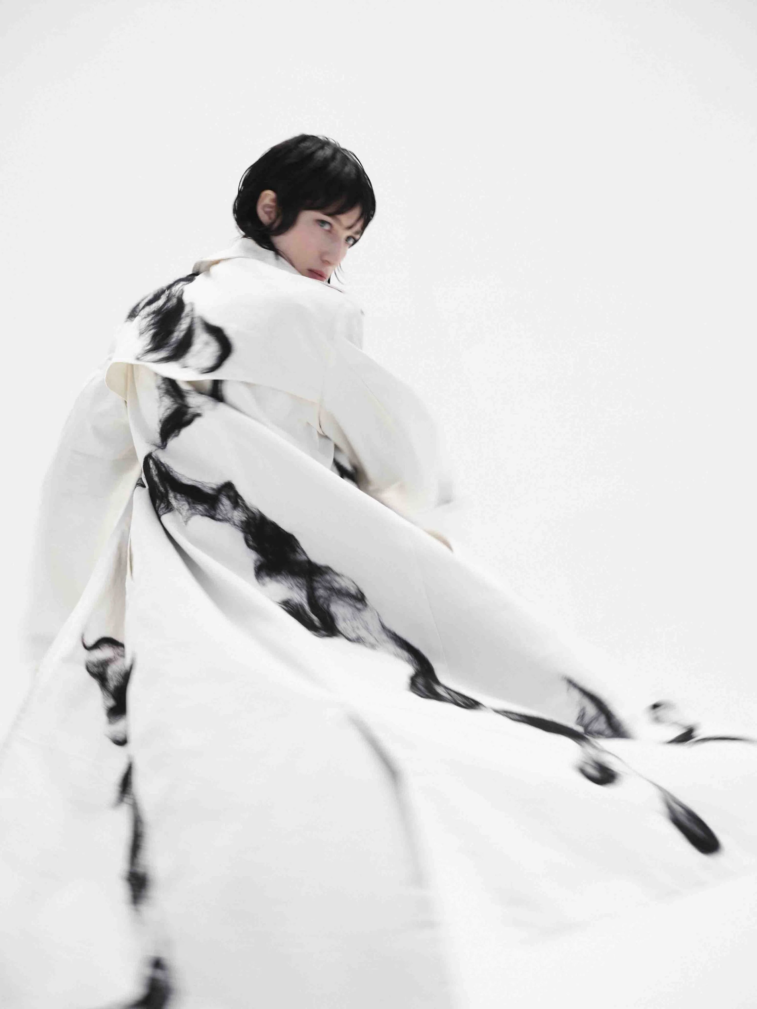 Person wearing a flowing white coat with black abstract patterns, turning to the side against a plain background.