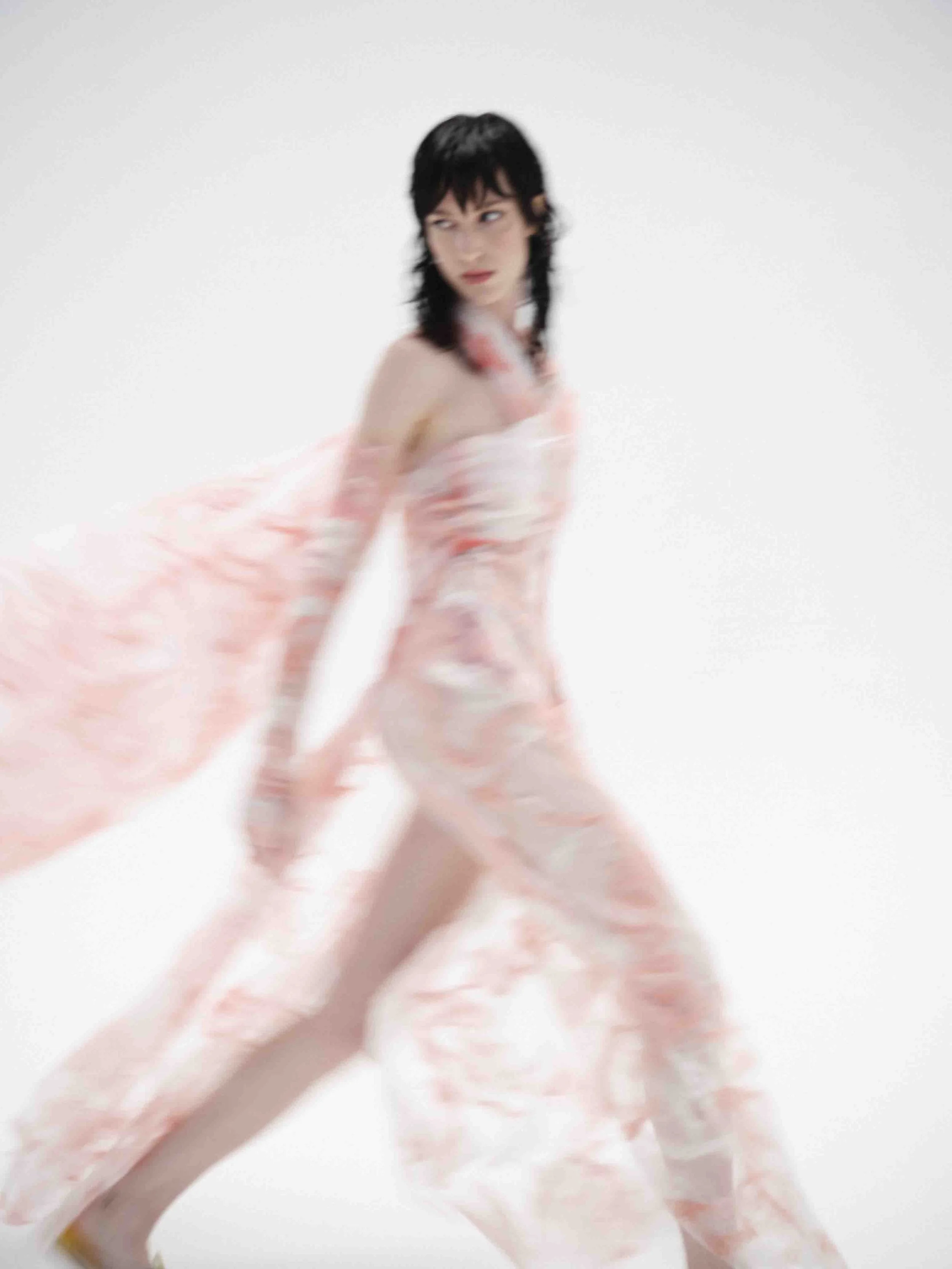 Blurred image of a person in a flowing, translucent pink dress walking.