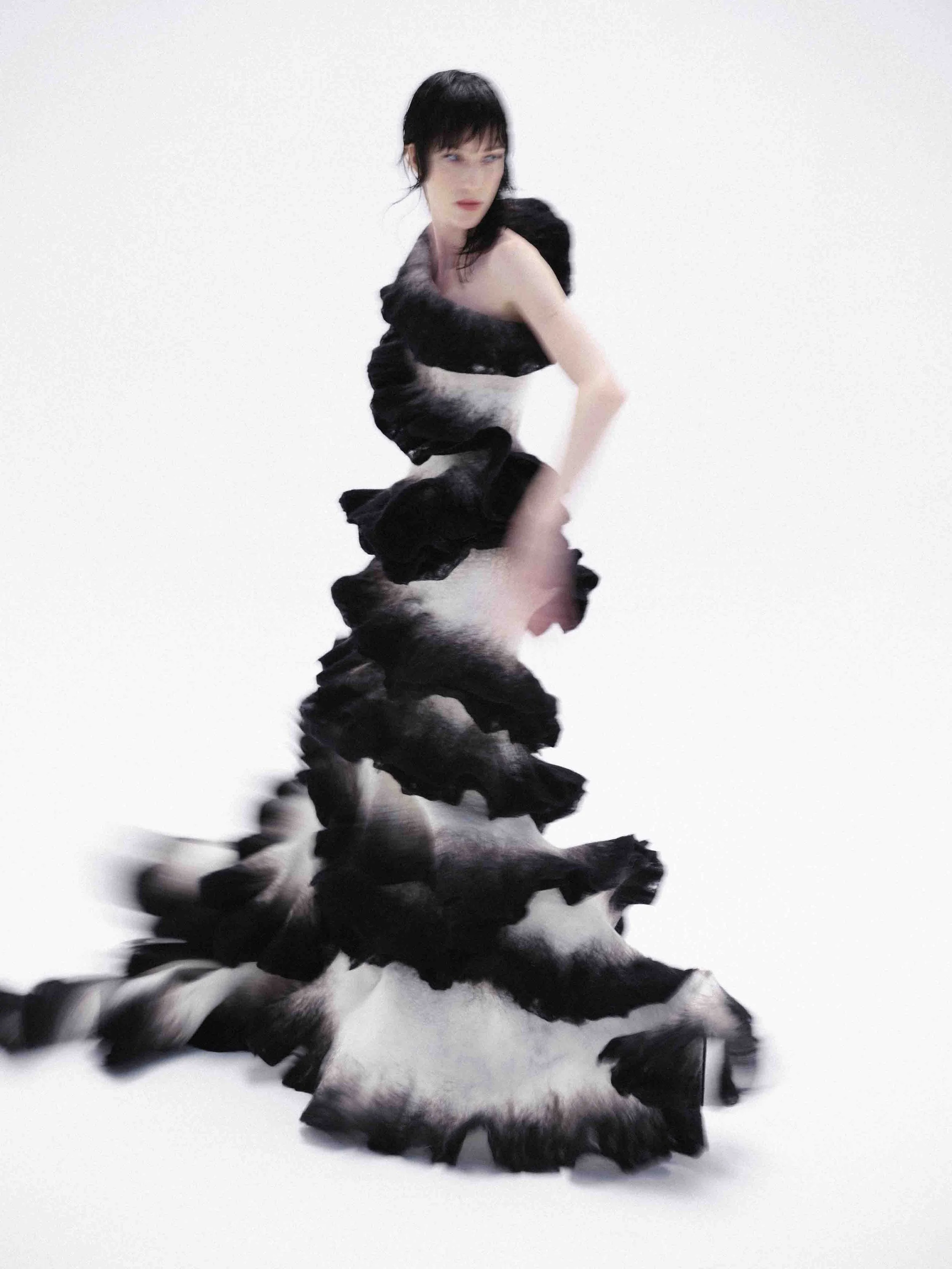 A person wearing a flowing, ruffled black and white gown stands in motion against a white background.