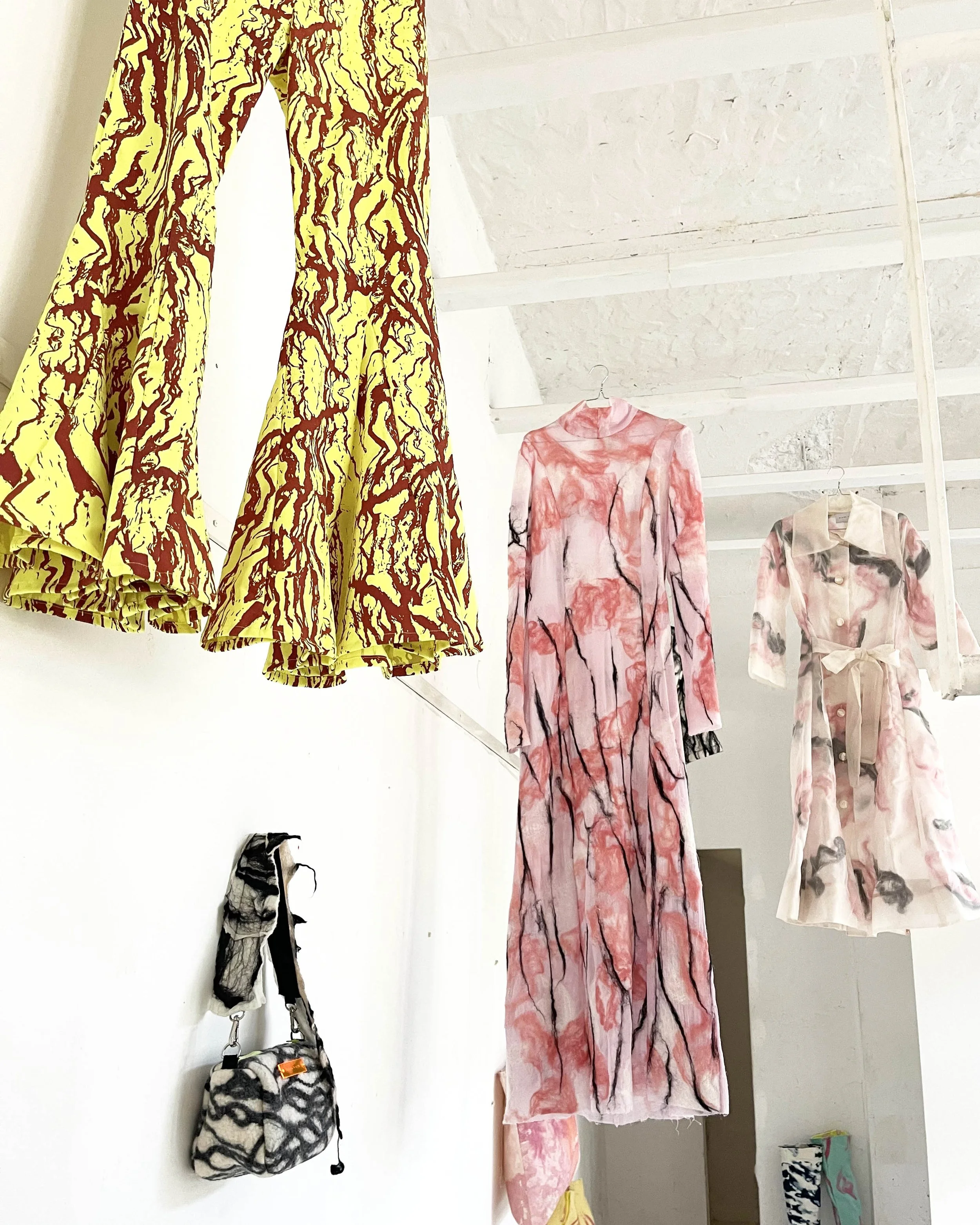 Colorful designer dresses and handbags displayed on the wall in a fashion store. The dresses have abstract, watercolor-like patterns in shades of red, pink, and beige. The handbags feature black and white patterns with some bright accents, hanging from hooks beneath the dresses.