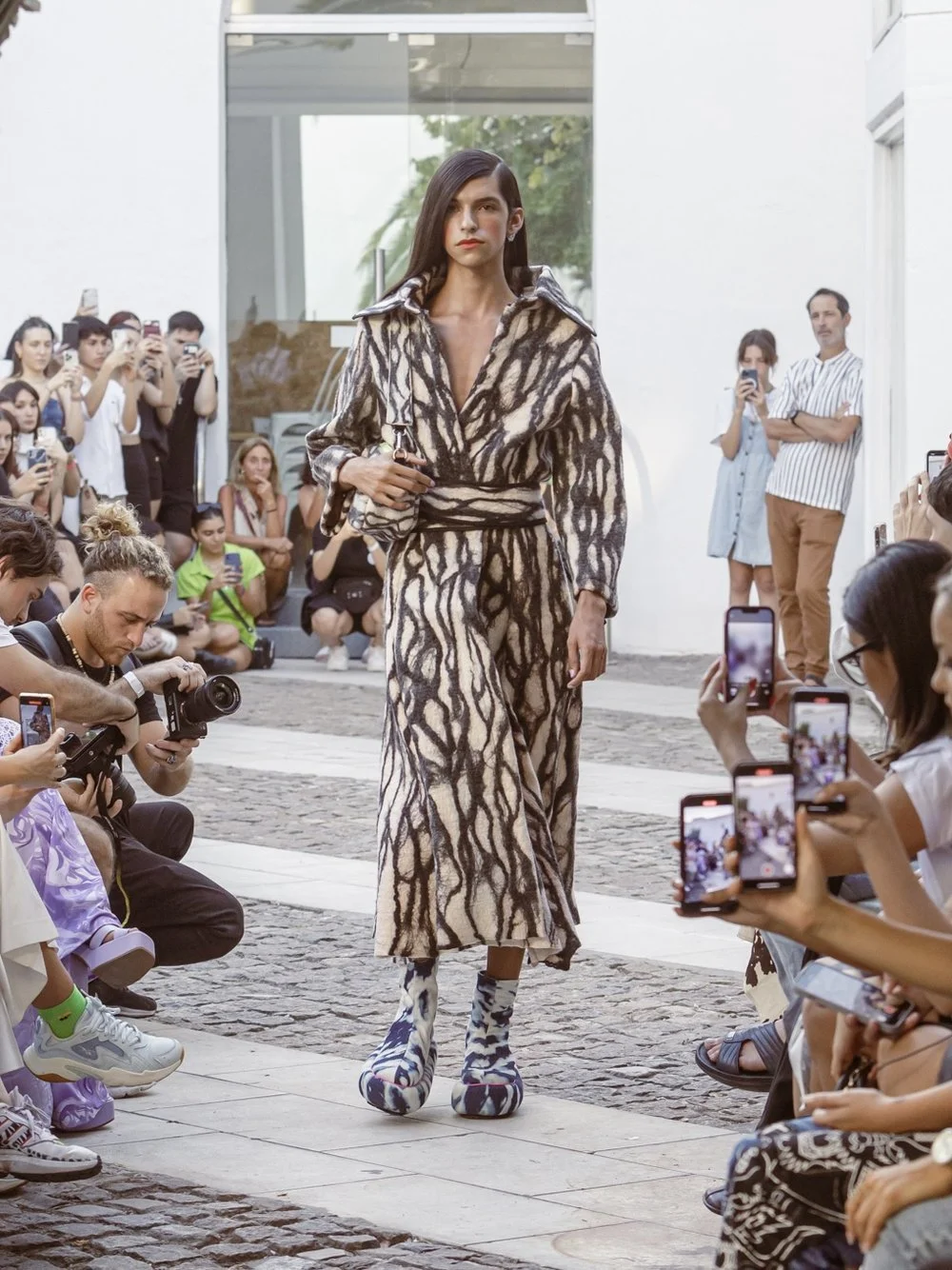 A fashion model walking down a runway during a fashion show, wearing a patterned robe-like outfit and distinctive shoes. The audience is taking photos using smartphones and cameras.