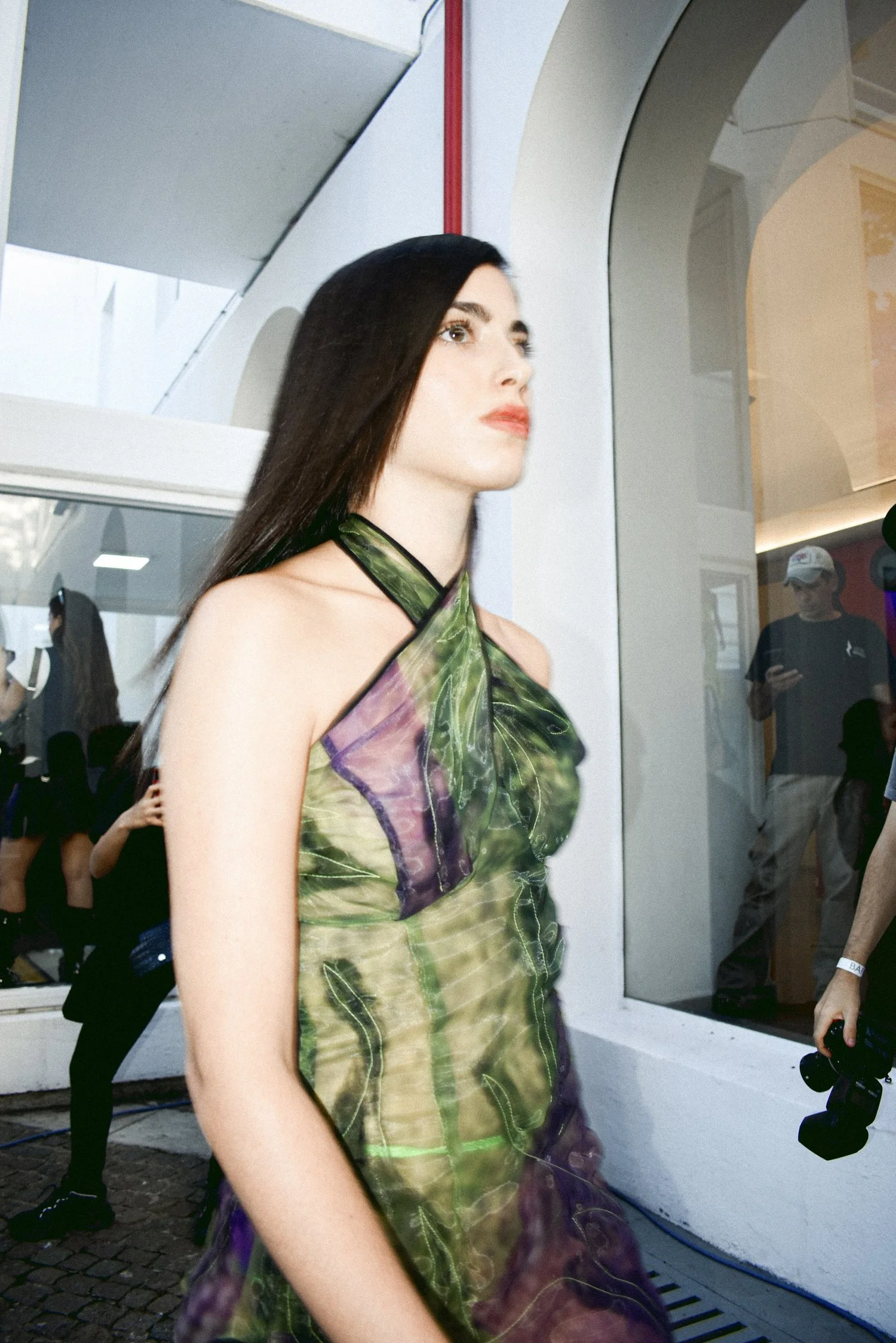 Person wearing a green and purple sheer dress walking in front of a building, with people taking photos in the background.