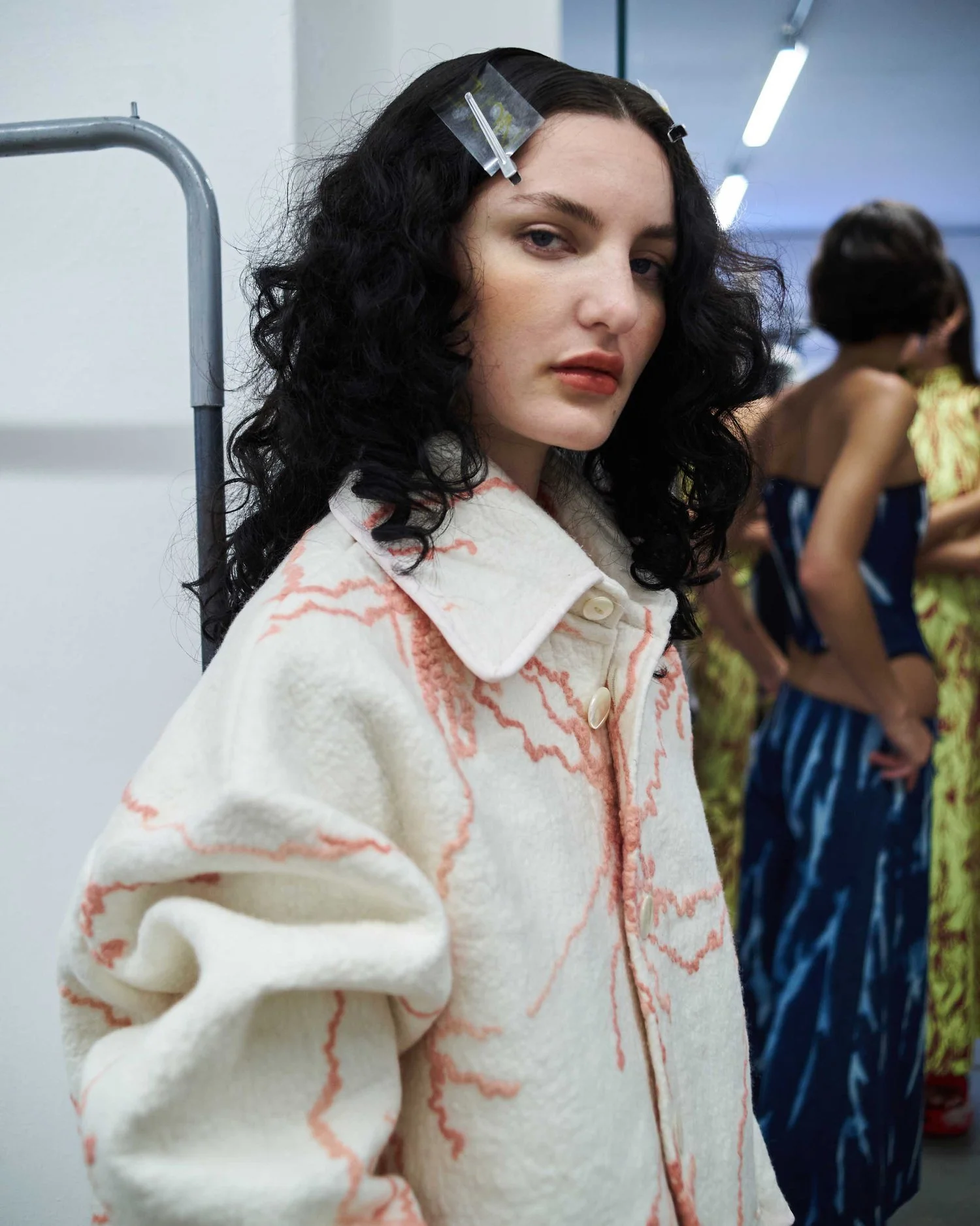 Fashion model with dark curly hair and hair clips wearing a white and pink textured jacket, posing indoors with blurred figures in colorful dresses in the background.