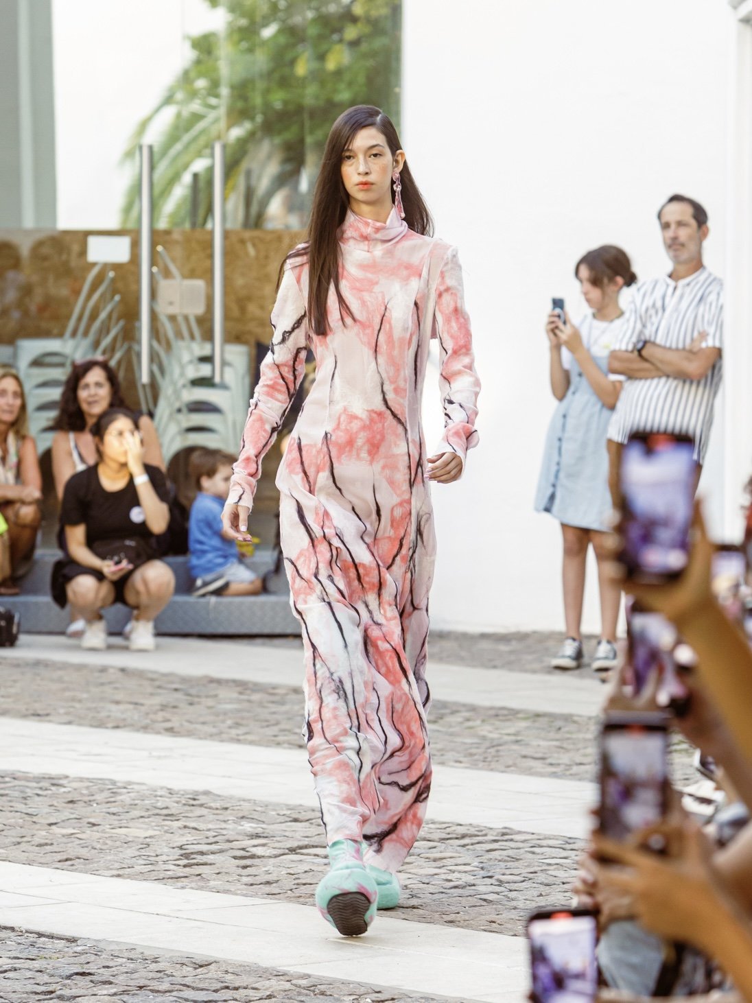 Model walking on a runway wearing a pink and white patterned outfit with blue-green shoes, audience taking photos.