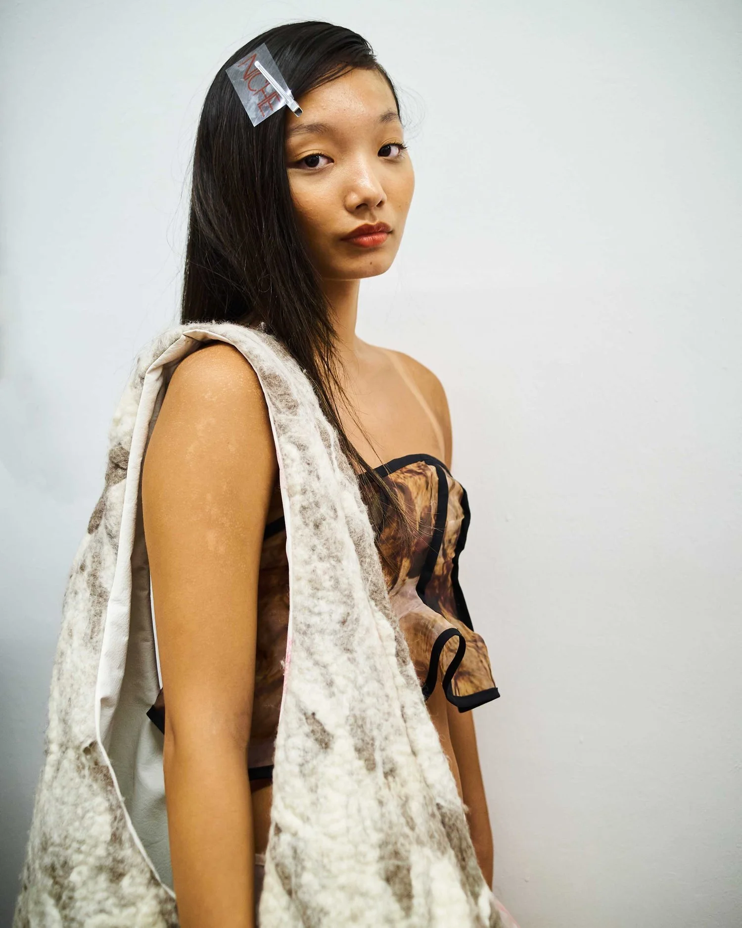 A woman with long hair and a hairpin wearing a textured, sleeveless top and furry scarf against a plain background.