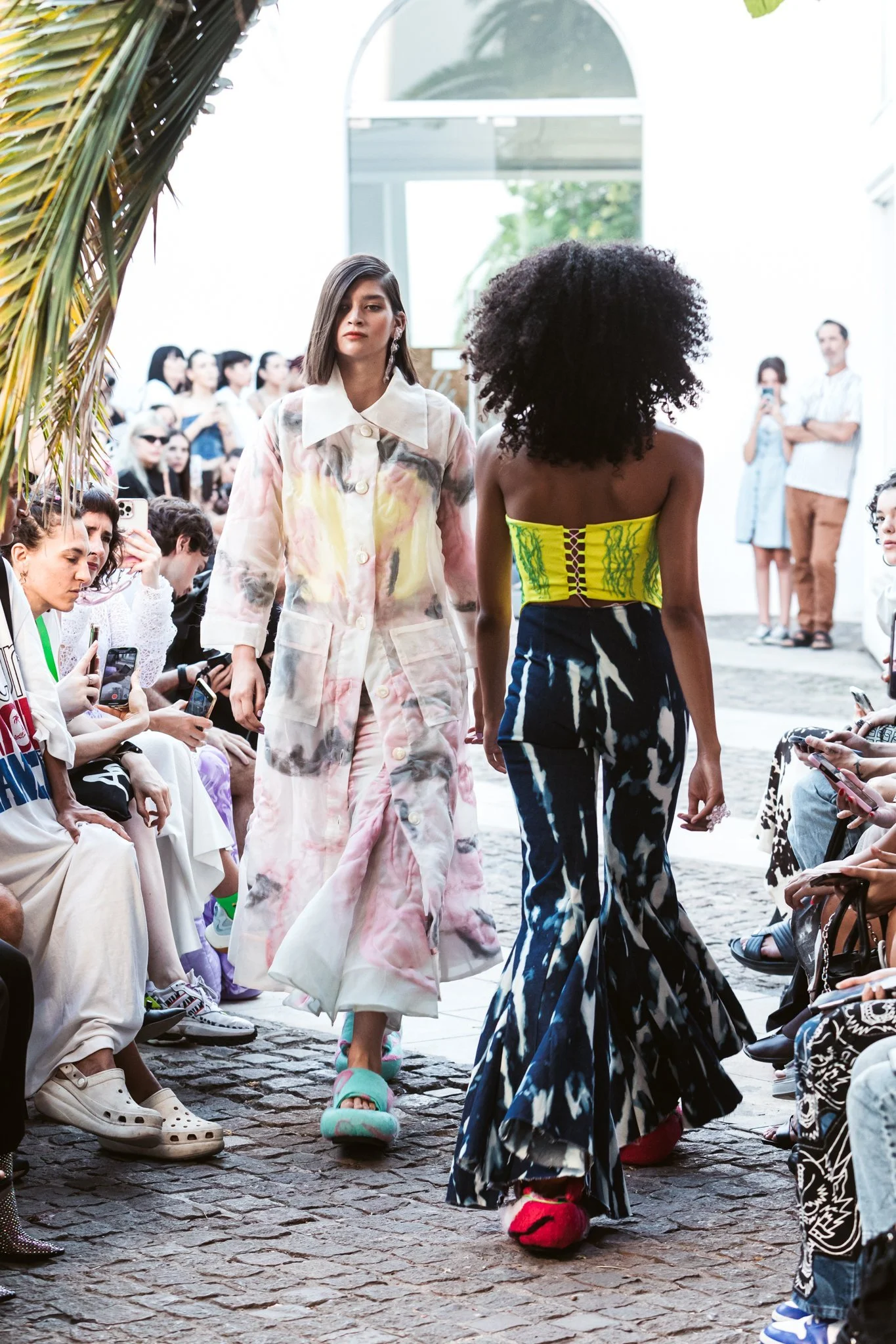 Fashion show with models walking the runway, one wearing a floral coat and the other in a colorful outfit, surrounded by seated audience.