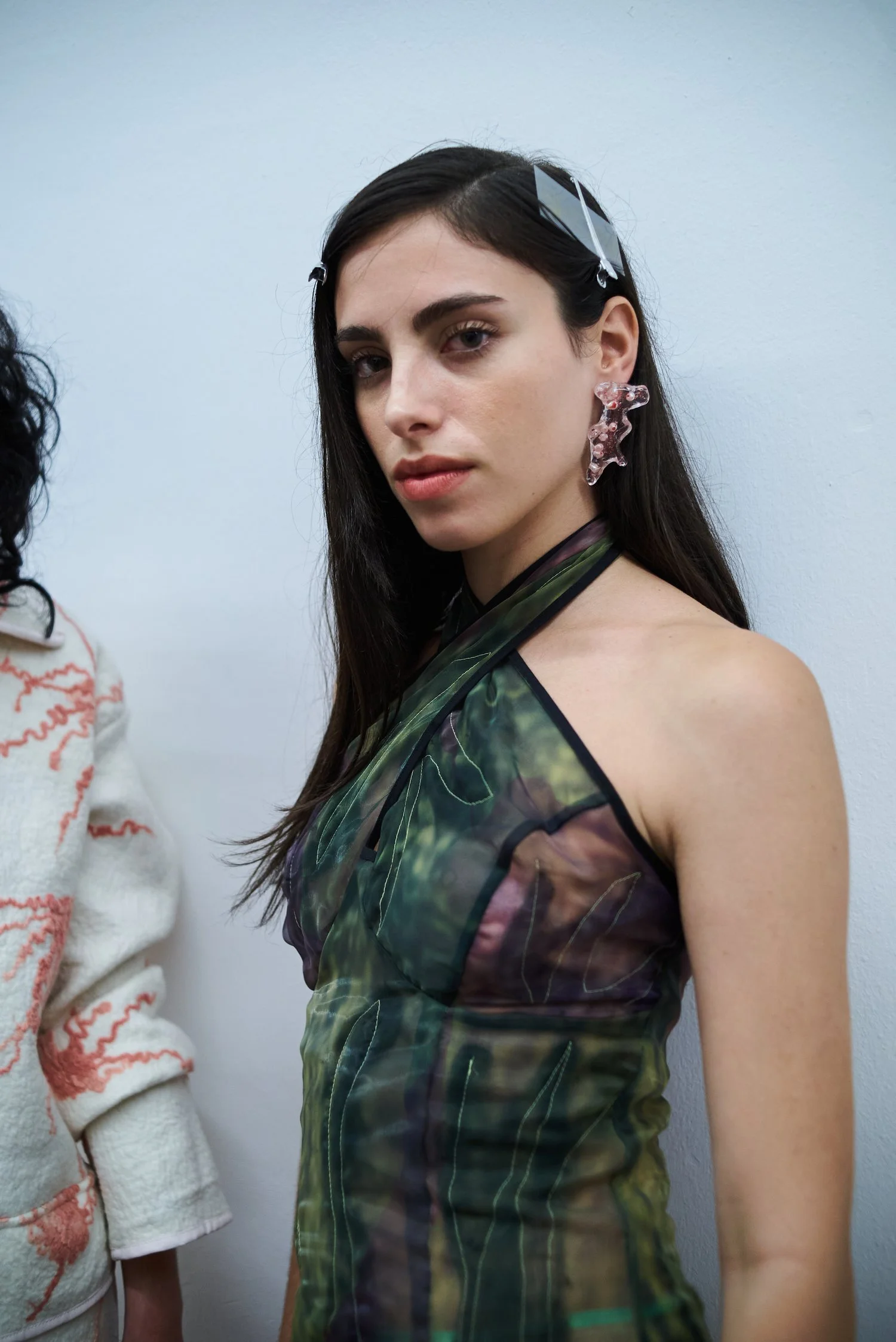 Fashion model backstage, wearing a green sheer top with pink earrings, and long dark hair with barrettes.