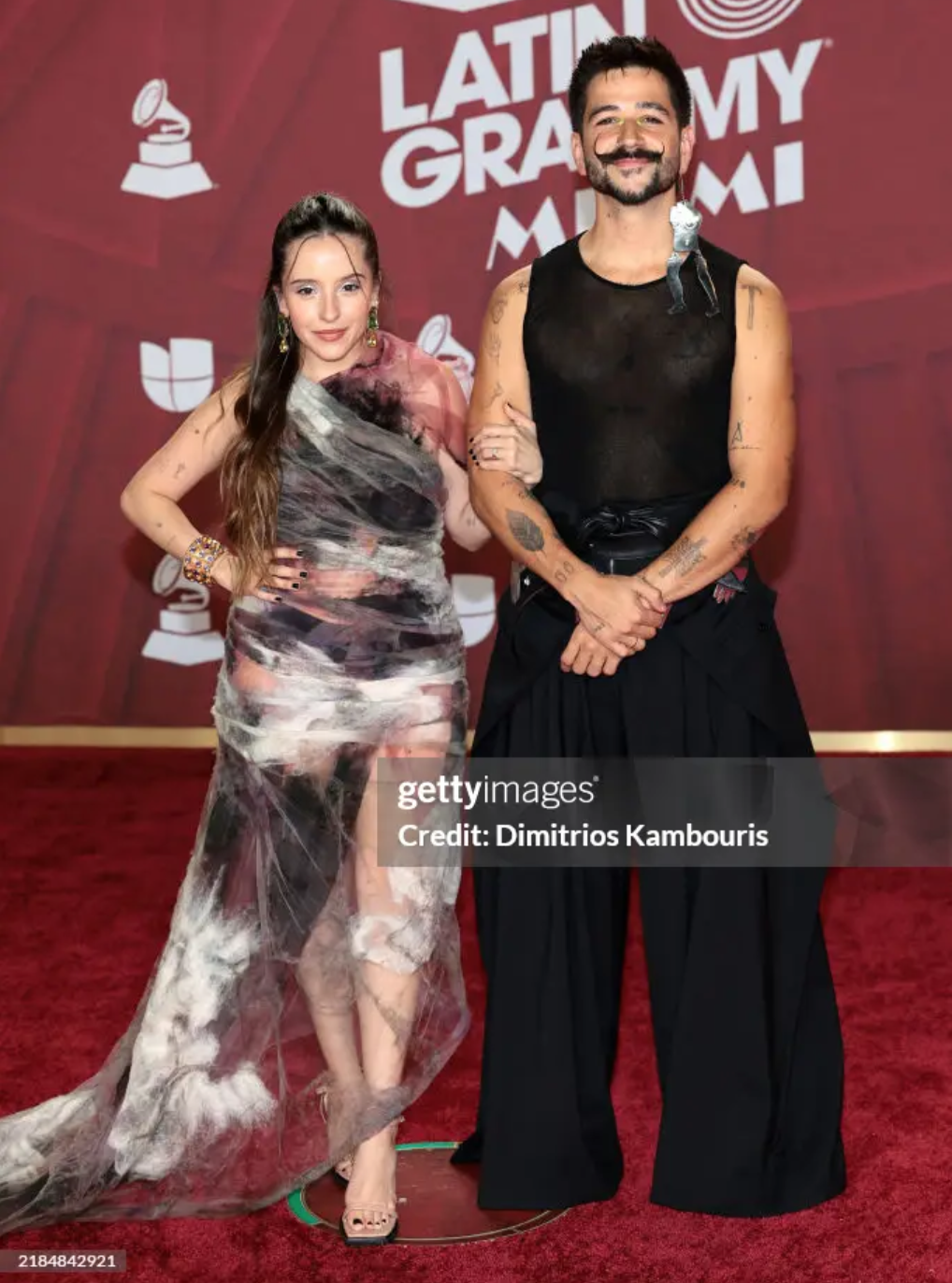 Two people standing on a red carpet at the Latin Grammy Awards, with a red backdrop behind them. The woman on the left has long brown hair and is wearing a sheer, multicolored dress with a slit, and the man on the right has short dark hair, a beard, 