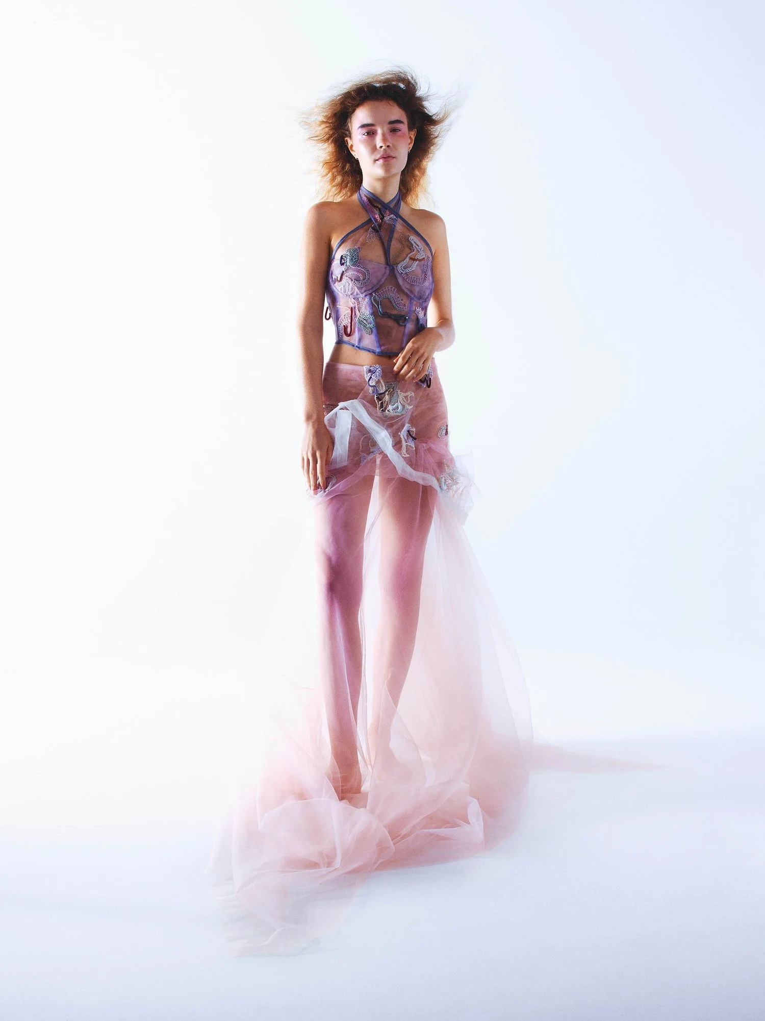 A woman wearing a sheer, flowing pink and purple outfit, posing against a light background.