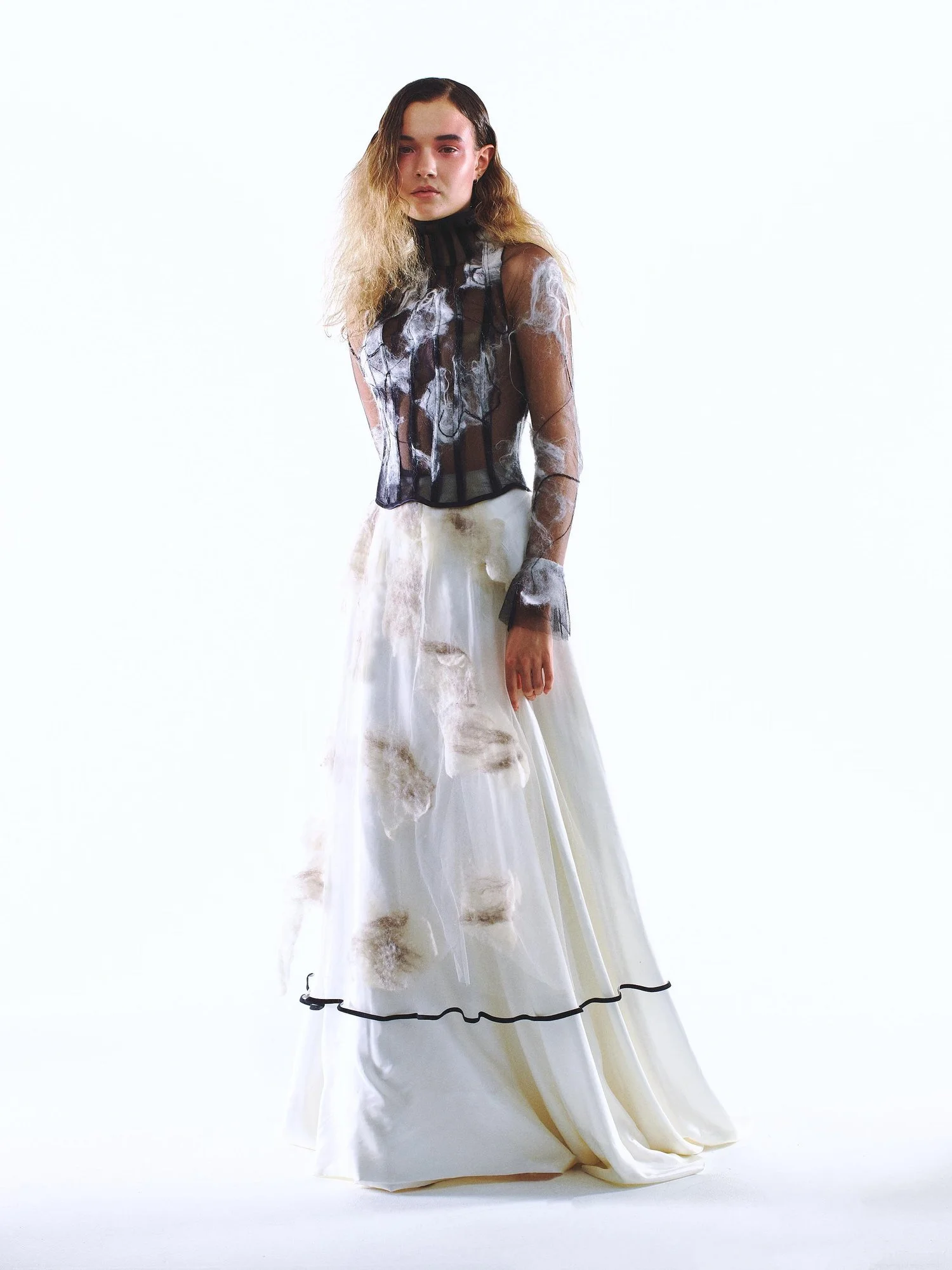 Woman wearing a sheer floral top and long white skirt with floral details, standing against a plain white background.